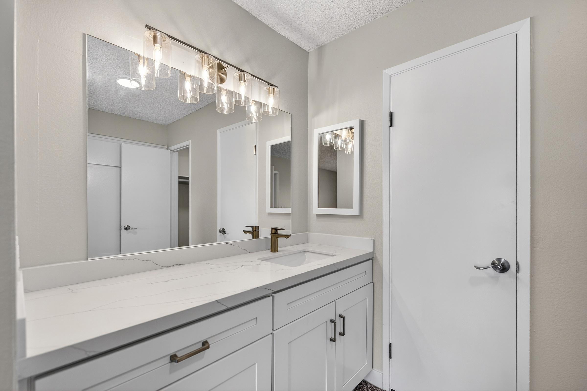 A modern bathroom featuring a white marble countertop with a sink, a wall-mounted mirror above, and stylish light fixtures. The walls are painted in a soft gray, and there is a closed white door on the right. The space is well-lit and clean, providing a contemporary aesthetic.
