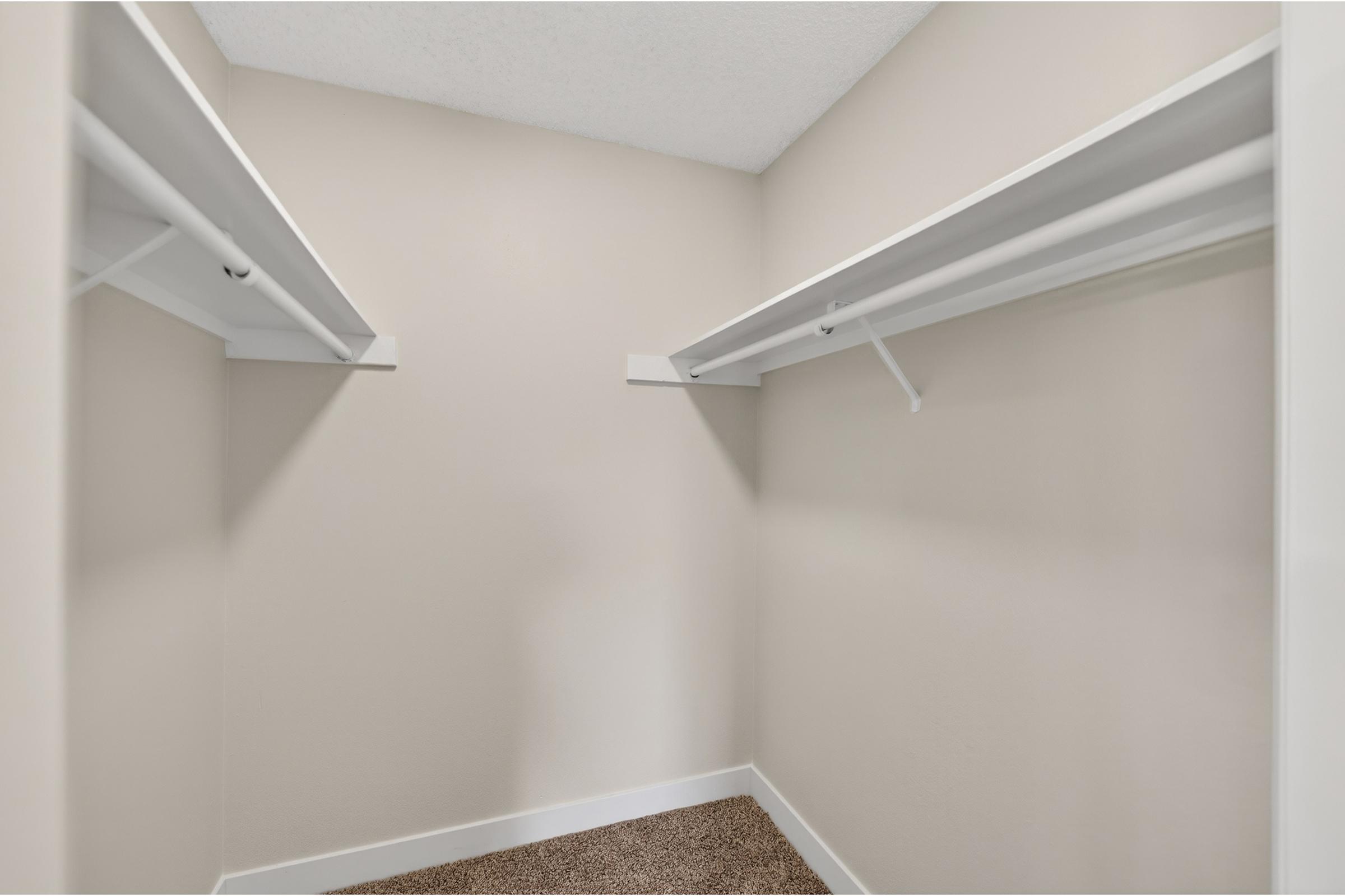 An empty closet with two white shelving units on opposite walls, featuring a neutral beige color scheme. The floor is carpeted in a light brown shade, and the space is well-lit, creating a clean and organized appearance.
