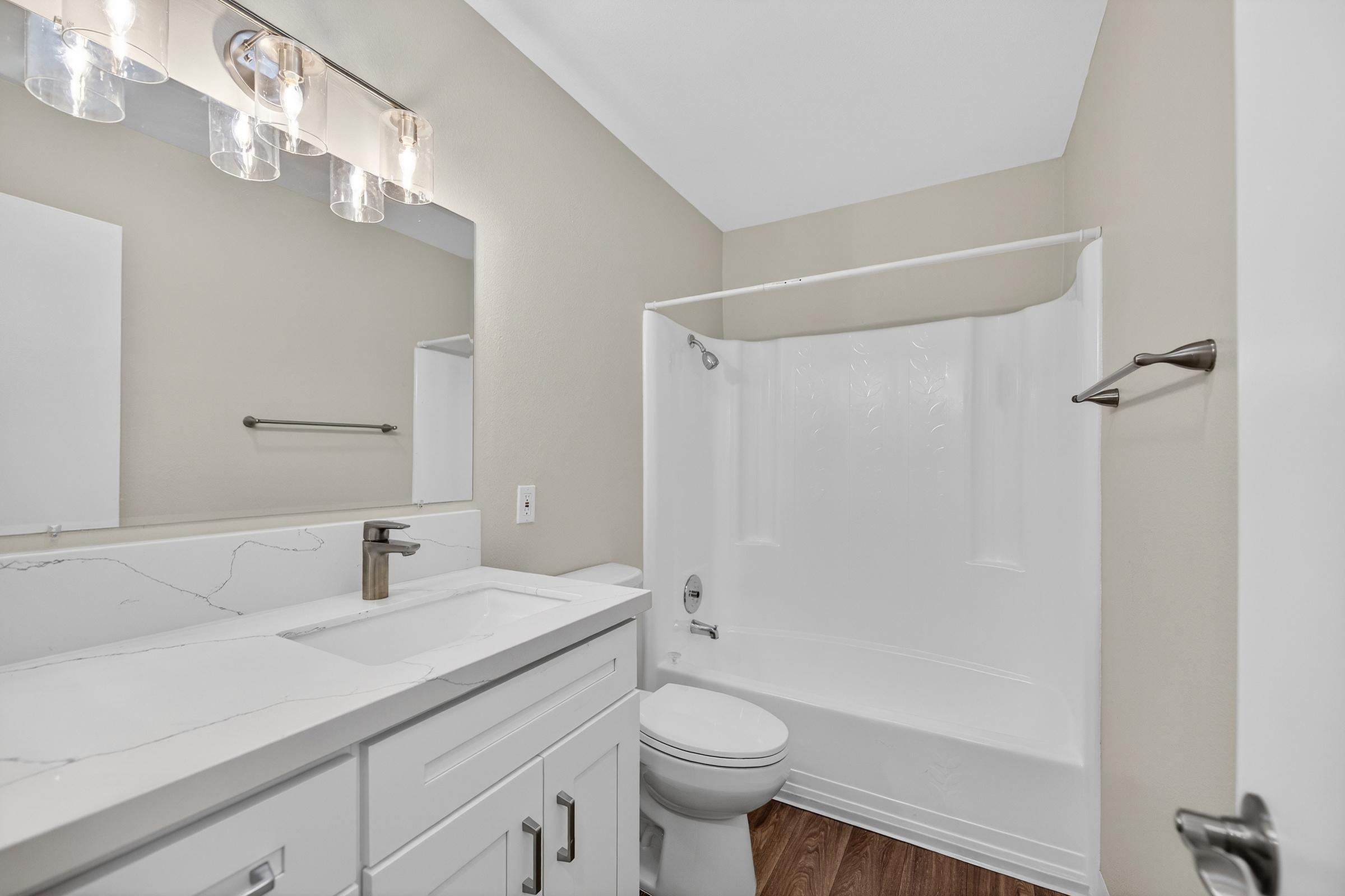 Bright and modern bathroom featuring a white bathtub with a shower, sleek white vanity with a rectangular sink, and a large mirror above the sink. The walls are painted in a neutral tone, and the flooring is a warm wood laminate, creating a clean and inviting atmosphere.