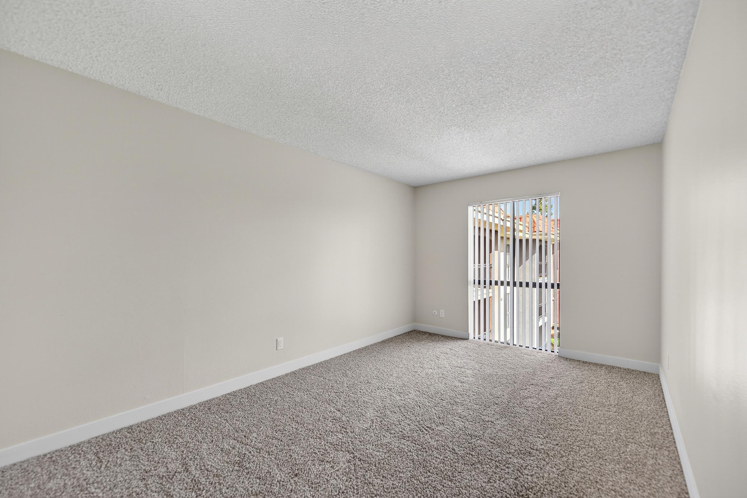 A spacious, empty room with light-colored walls and a textured ceiling. The floor is covered in soft, gray carpeting. A set of vertical blinds partially covers a window, revealing a view of an outdoor area. The overall ambiance is bright and inviting, perfect for personalizing with furniture and decor.