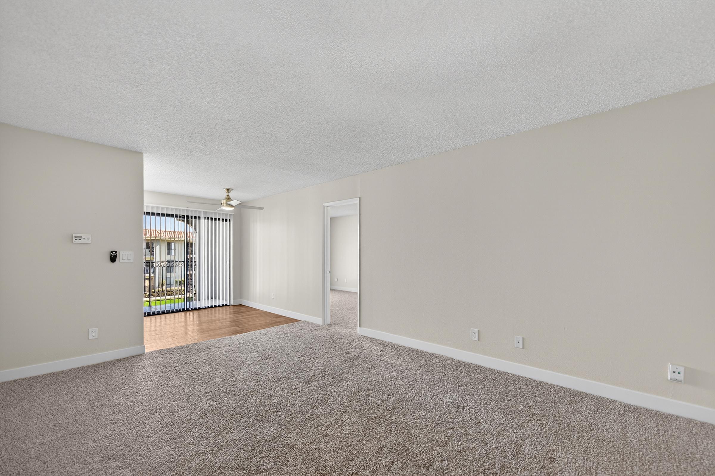 Spacious interior of a living area with neutral walls and carpet. A door leads to an outdoor area with vertical blinds. Natural light enters from the sliding glass door, showcasing the open layout. The ceiling features a ceiling fan, adding to the room's comfort.