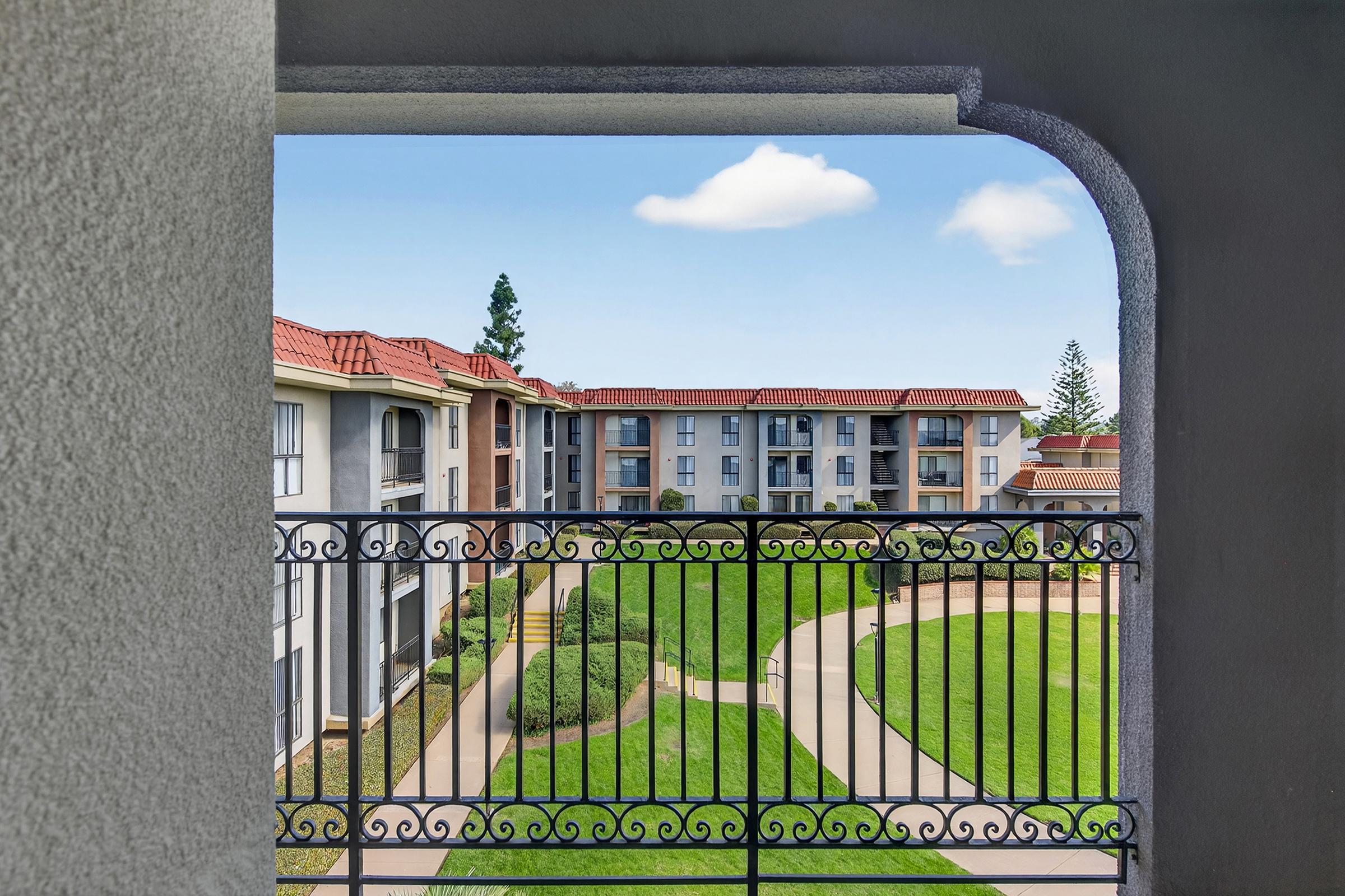 View from a balcony overlooking a landscaped courtyard with green grass and walking paths, framed by an ornate railing. Several apartment buildings with red roofs are visible in the background under a clear blue sky.
