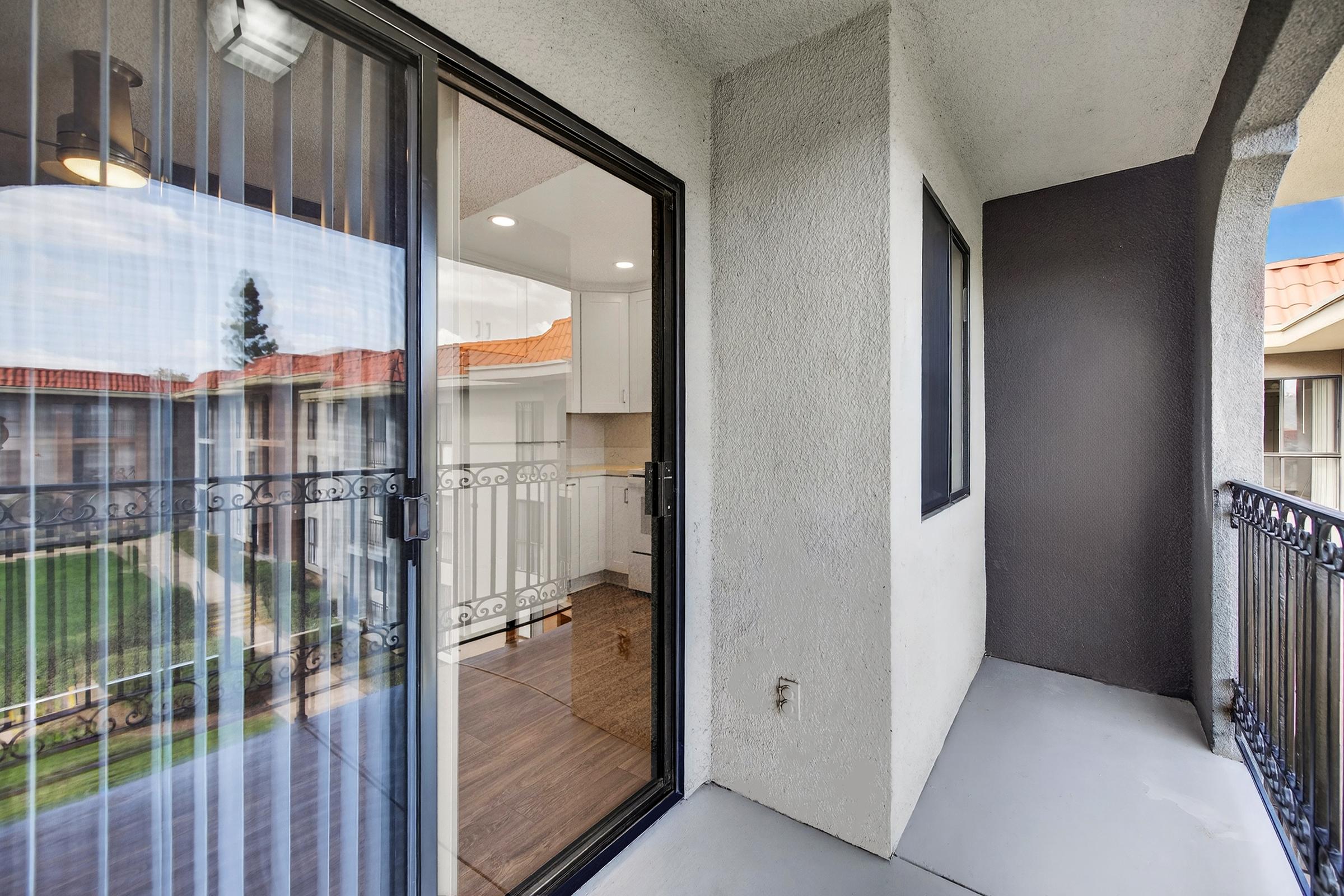View from a balcony looking into an apartment with large sliding glass doors. The area features wooden flooring inside and a railing outside. The backdrop includes other buildings and a small grassy area, with trees visible in the distance under a clear sky.