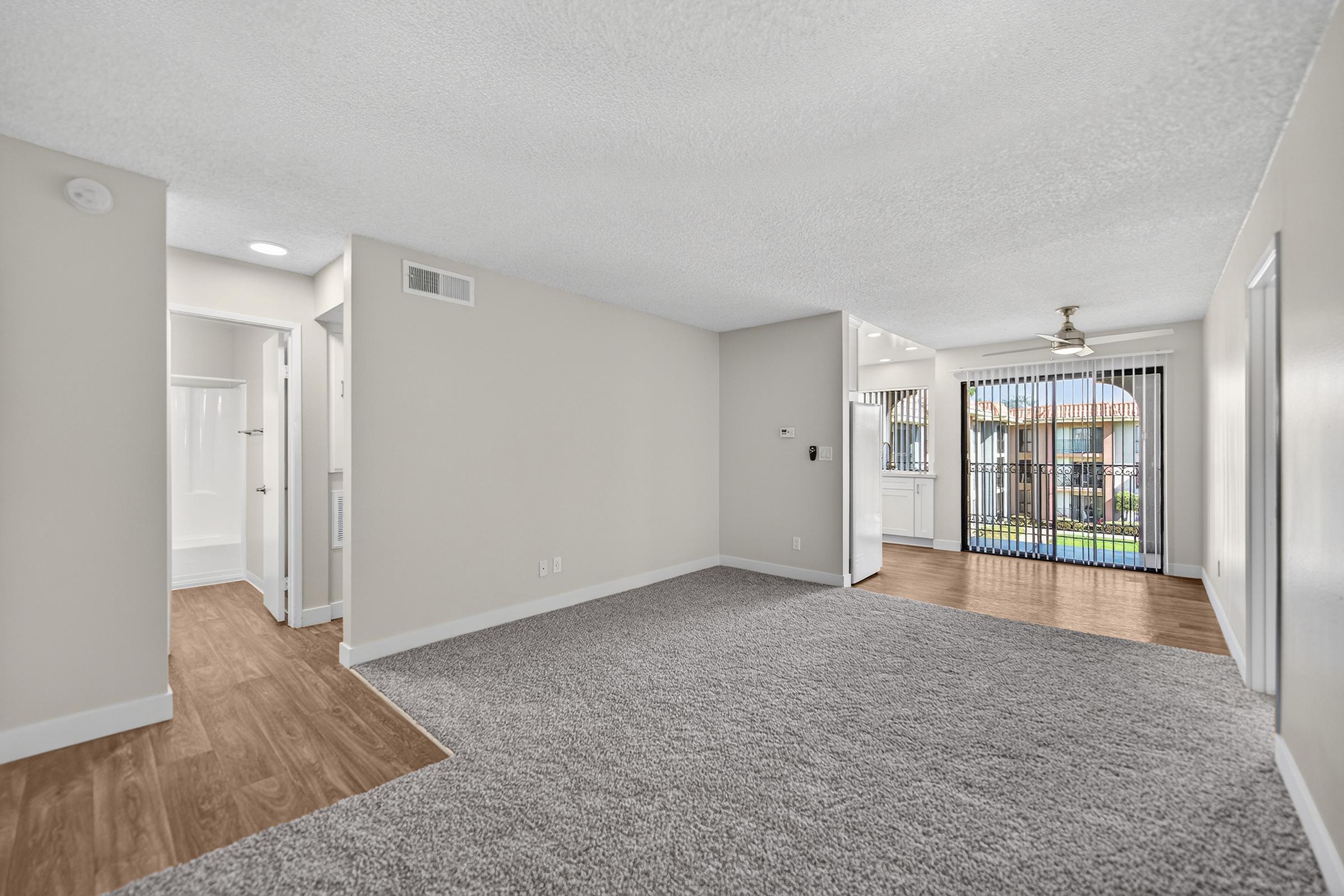 A spacious interior view of a light-colored room featuring plush carpeting, a ceiling fan, and large windows with vertical blinds. There’s an adjoining area with a bathroom and a corridor leading to another room. The flooring transitions from carpet to a wooden finish, creating an inviting atmosphere.
