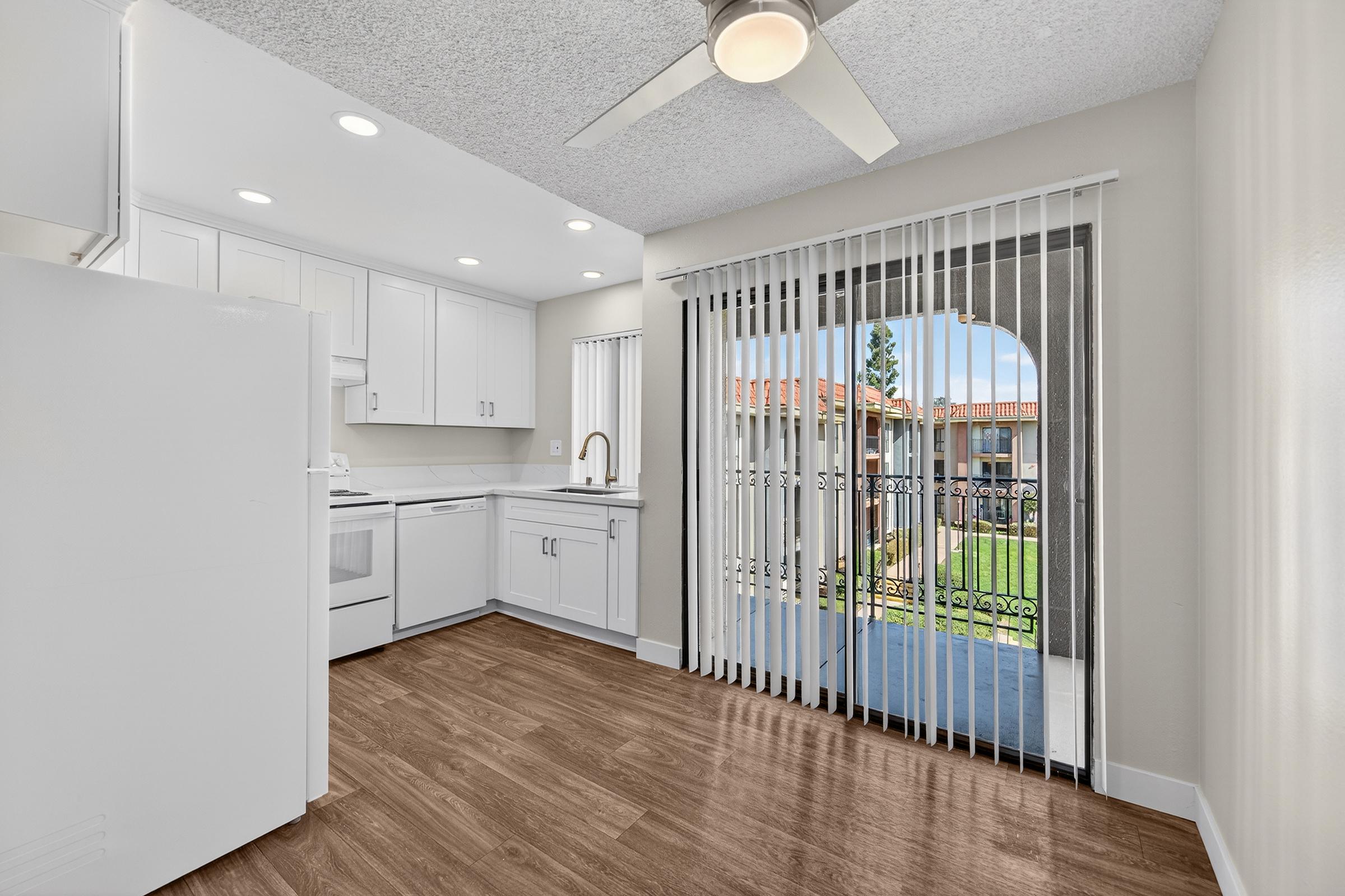 A modern kitchen with white cabinets and appliances, featuring a ceiling fan. There's a large window with vertical blinds that opens to a balcony with a view of greenery. The floor has a warm wood finish, and natural light brightens the room.