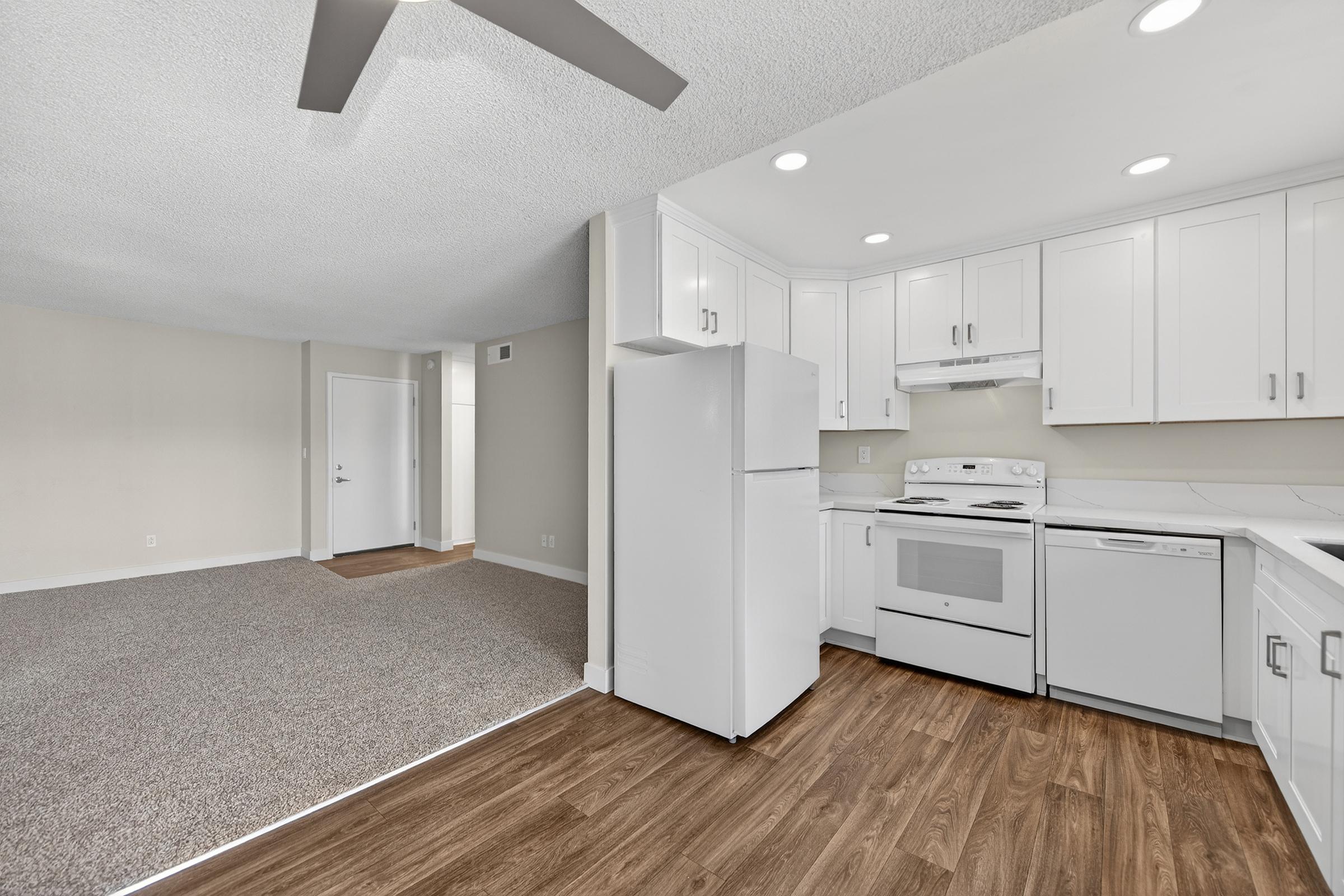Modern kitchen with white cabinetry and appliances, including a refrigerator and stove. Adjacent to the kitchen is a spacious living area with beige carpet and light walls, featuring a ceiling fan. An open doorway leads to another room, creating a bright and inviting atmosphere.