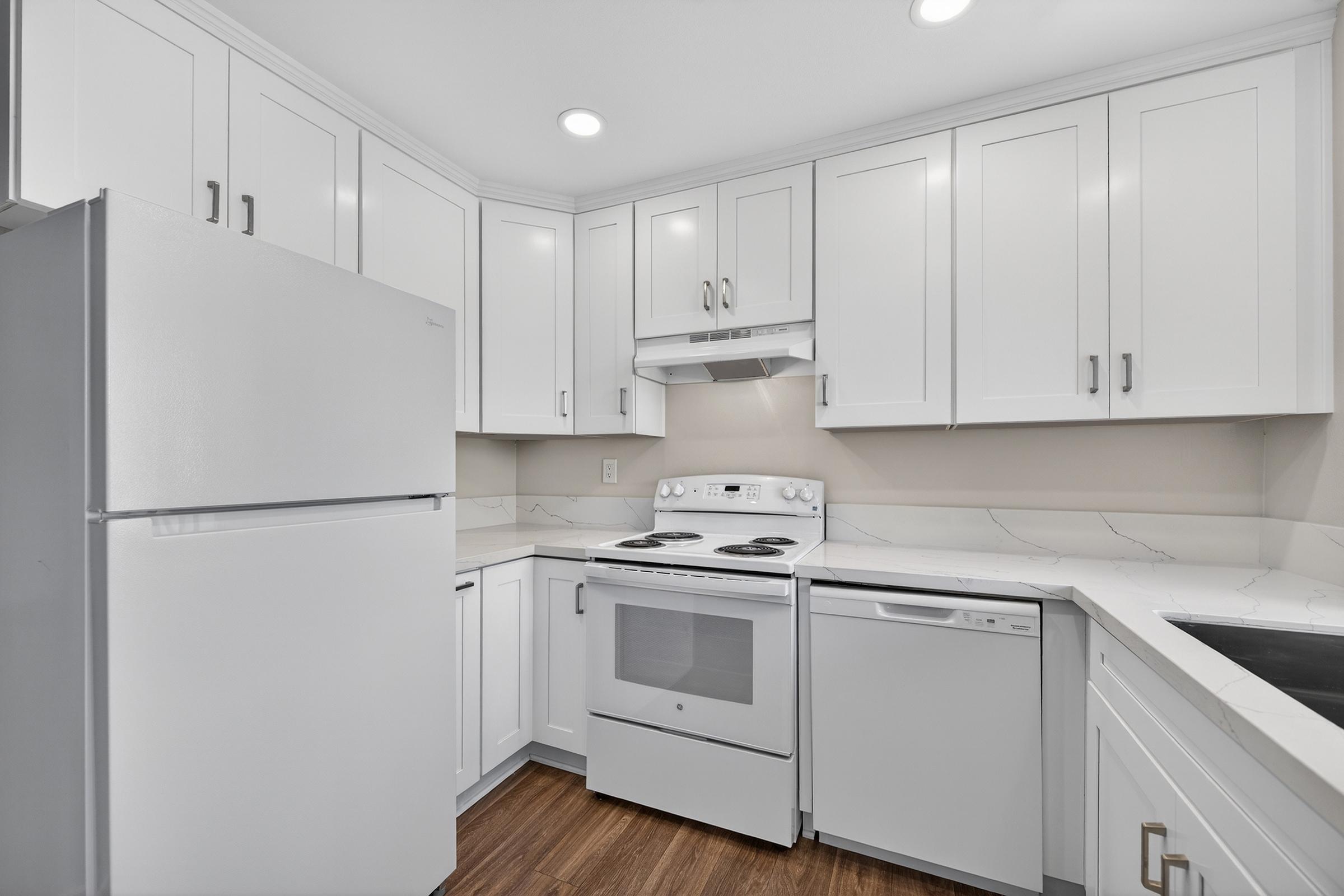 Modern kitchen featuring white cabinets, a white refrigerator, an oven, and a dishwasher. The countertops are light-colored with a subtle pattern, complemented by warm wood flooring. Recessed lighting illuminates the space, creating a clean and bright atmosphere.