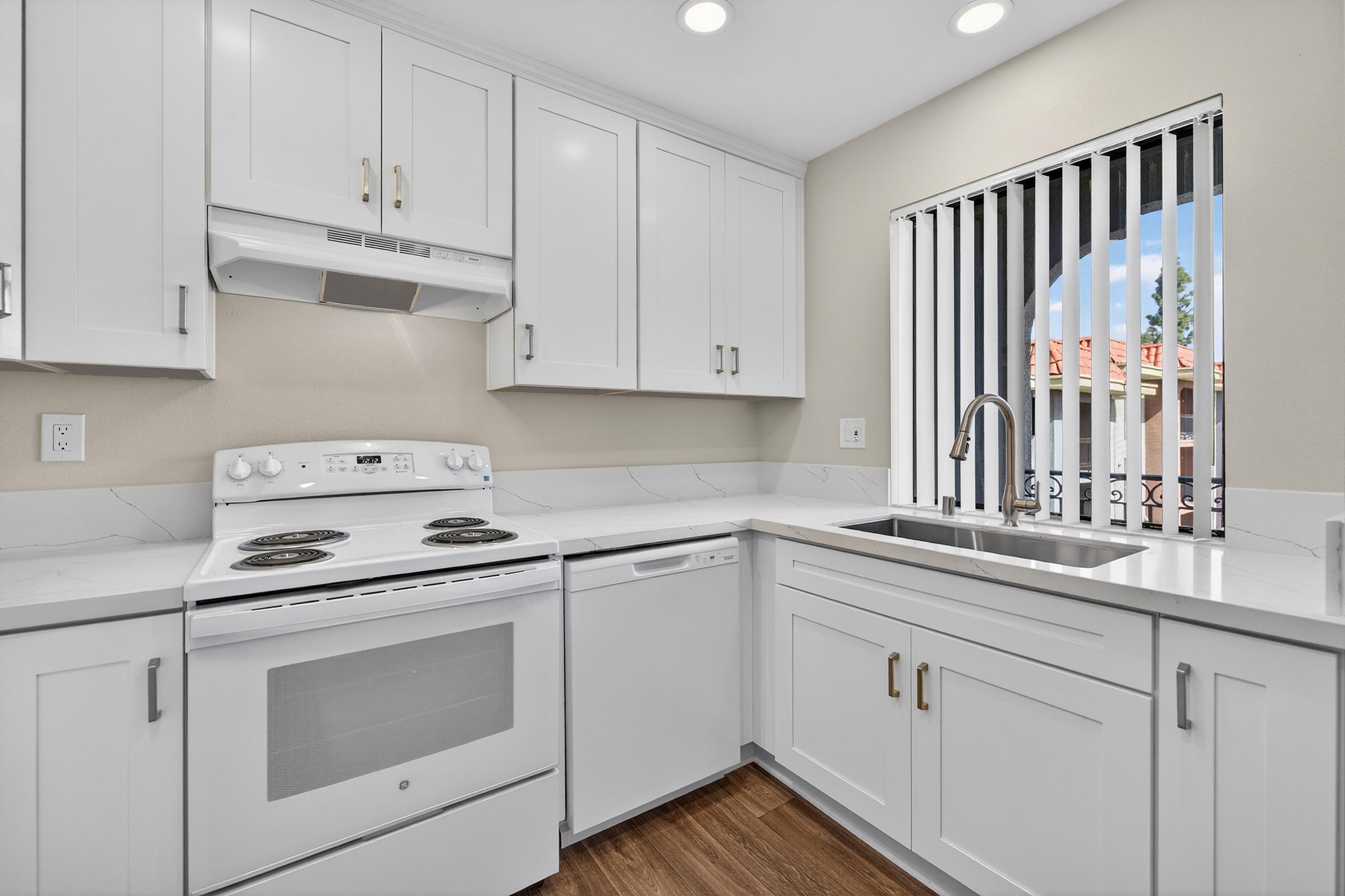 Modern kitchen featuring white cabinetry, a white stove and dishwasher, a stainless steel sink, and a window with vertical blinds. The countertop has a marble-like pattern, and there is a soft light coming from recessed ceiling fixtures. The space is well-organized and bright.