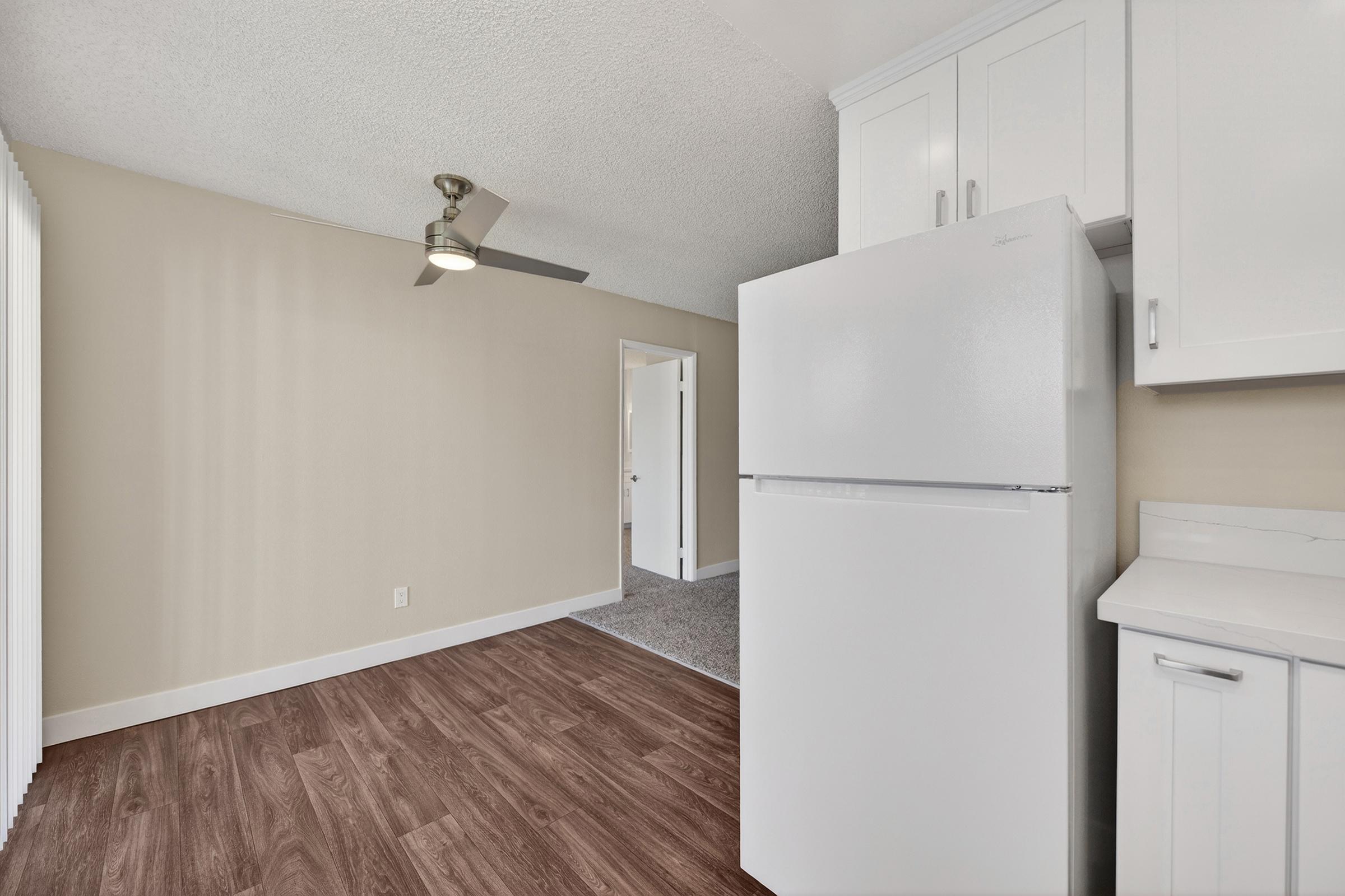 Bright and spacious kitchen area featuring a white refrigerator, modern cabinetry, wood-style flooring, and a ceiling fan. Light-colored walls and a doorway lead to another room, creating an inviting and fresh atmosphere.