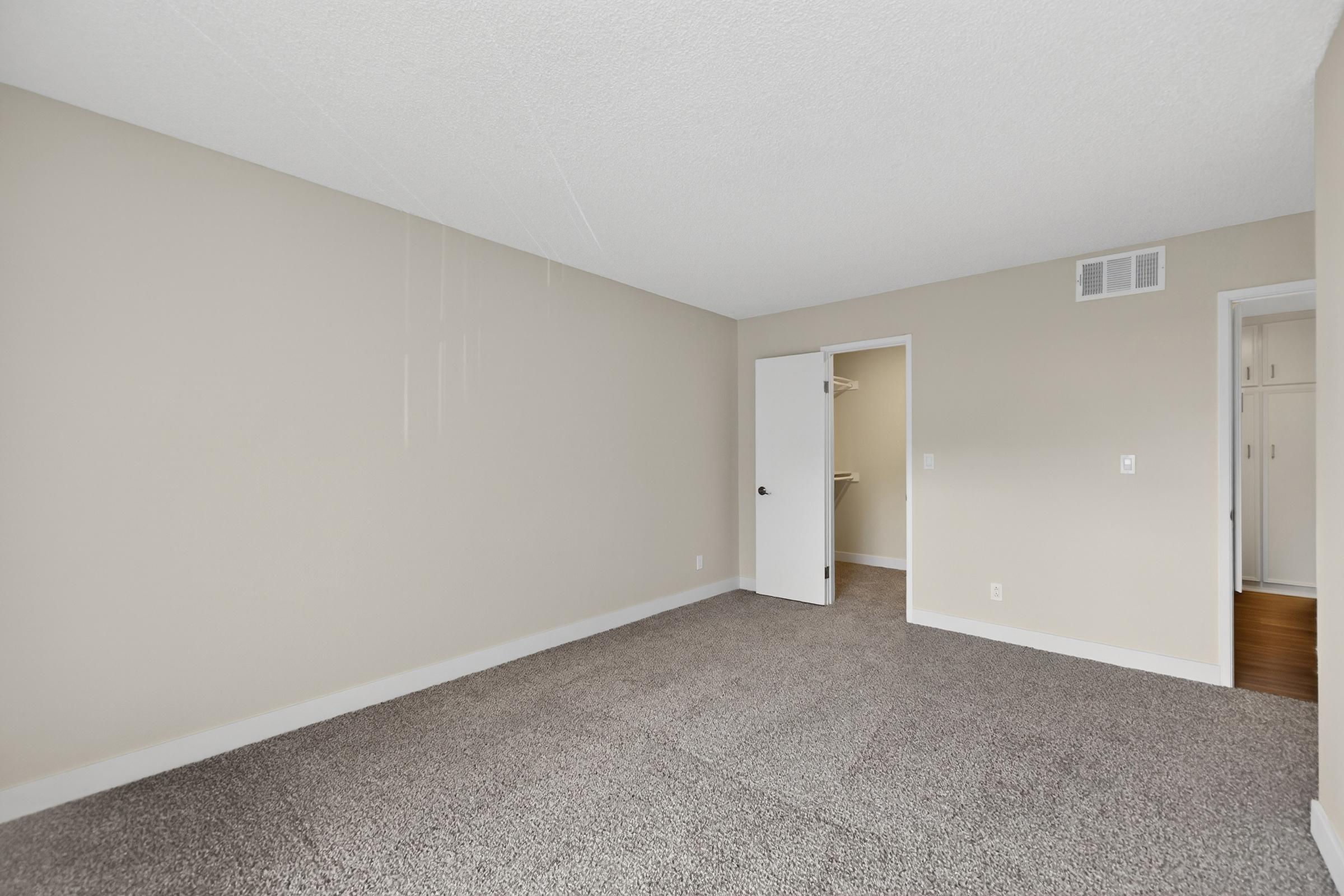 Empty room with light beige walls and carpeted flooring. A closed door leads to a closet on the right, while another door opens to a small room or storage area. The ceiling has a textured design, and the overall space is clean and uncluttered, suitable for various uses.