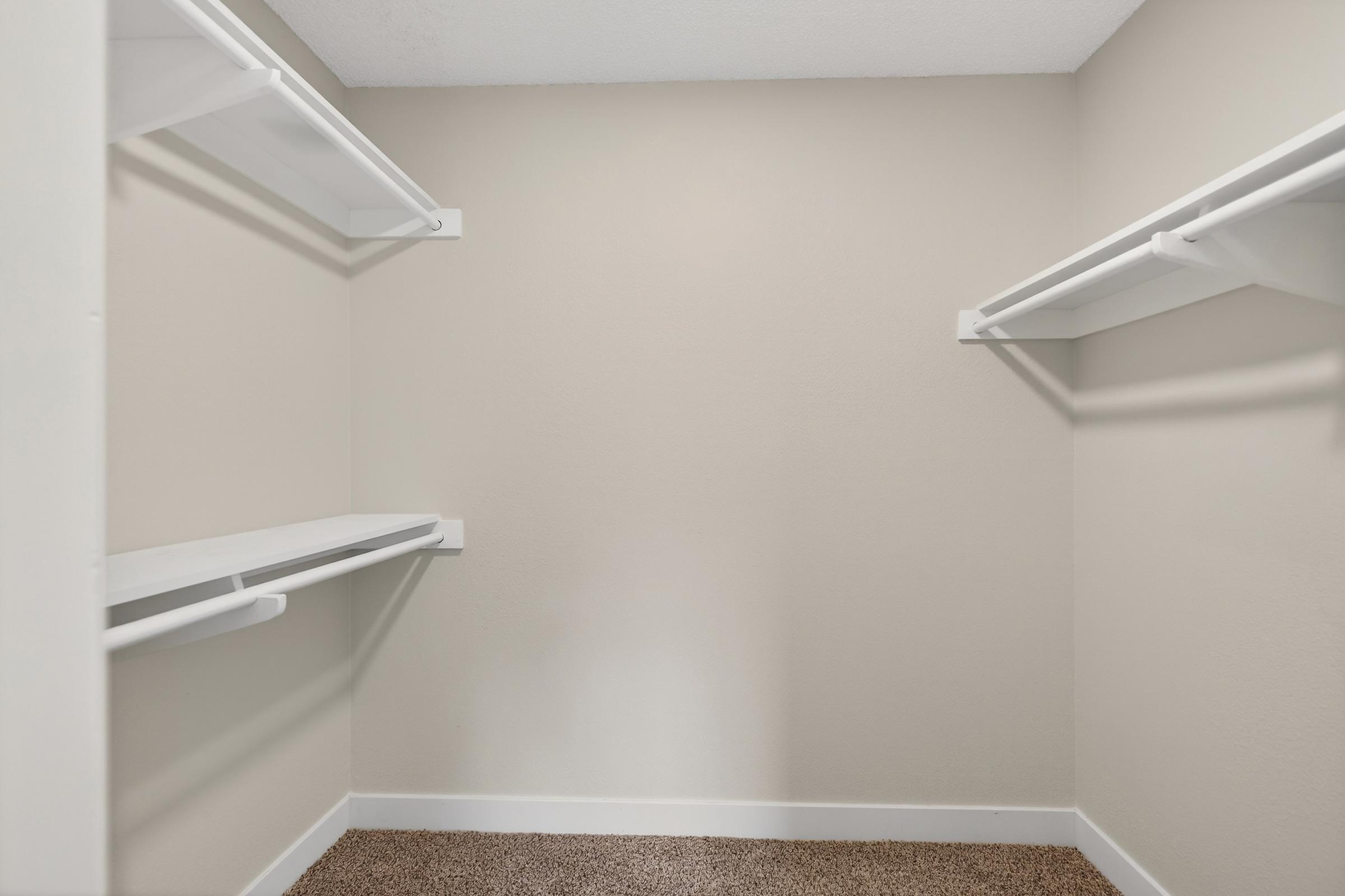 Empty closet with light-colored walls and two white shelves mounted on either side. The floor is covered in carpet, and there is a clear, open space in the center. The overall look is clean and minimalistic, suitable for storage or organization.