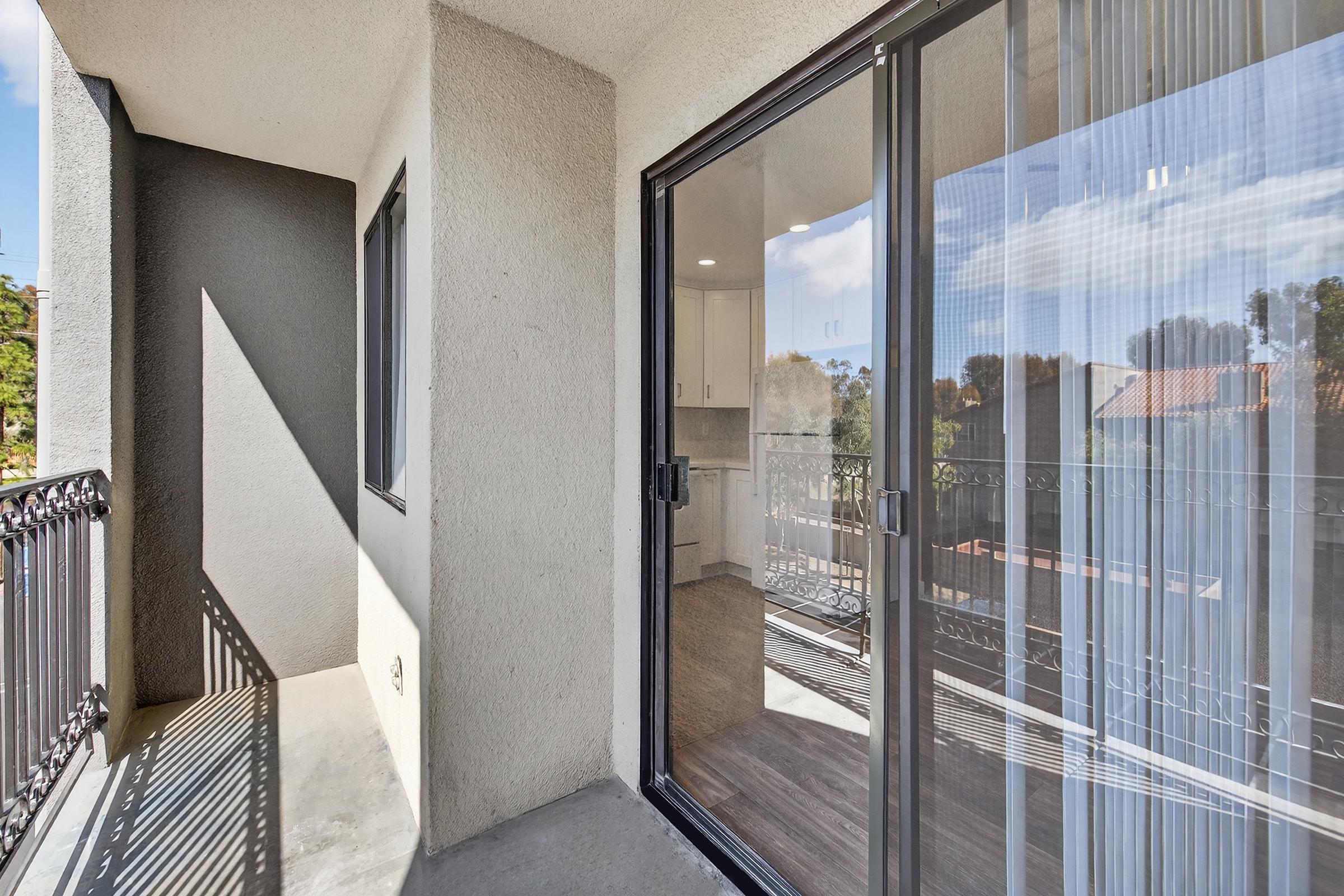 A view from a balcony showing a sliding glass door leading to an interior space. The balcony features a railing and is illuminated by natural light, with the exterior walls painted in neutral tones. In the background, there are glimpses of trees and rooftops under a blue sky with clouds.