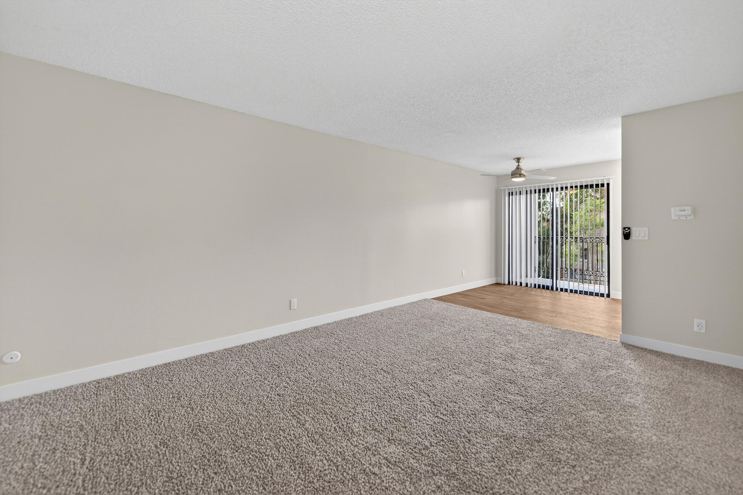 Spacious and bright living area featuring beige walls, light-colored carpet, and wood flooring. Large sliding glass door provides natural light and access to a balcony. A ceiling fan hangs from the textured ceiling, creating a modern and inviting atmosphere.