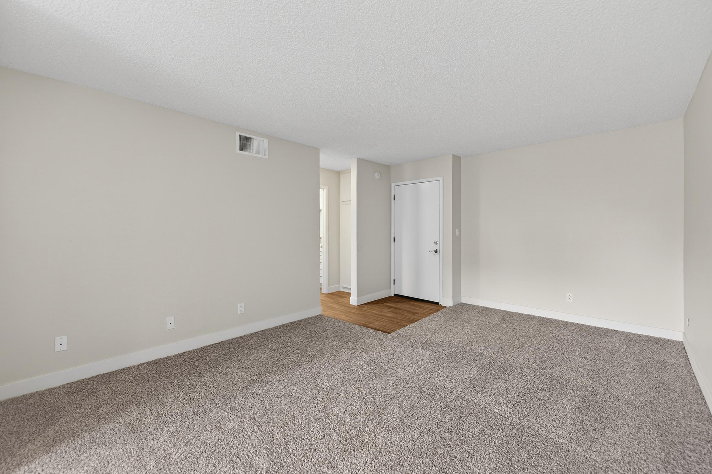 Spacious and empty room with light-colored walls and a textured ceiling. The floor is covered in plush carpeting, and there is a doorway leading to another room in the background. A white door is visible, adding to the bright and airy feel of the space. Ideal for personalized decoration and furnishing.