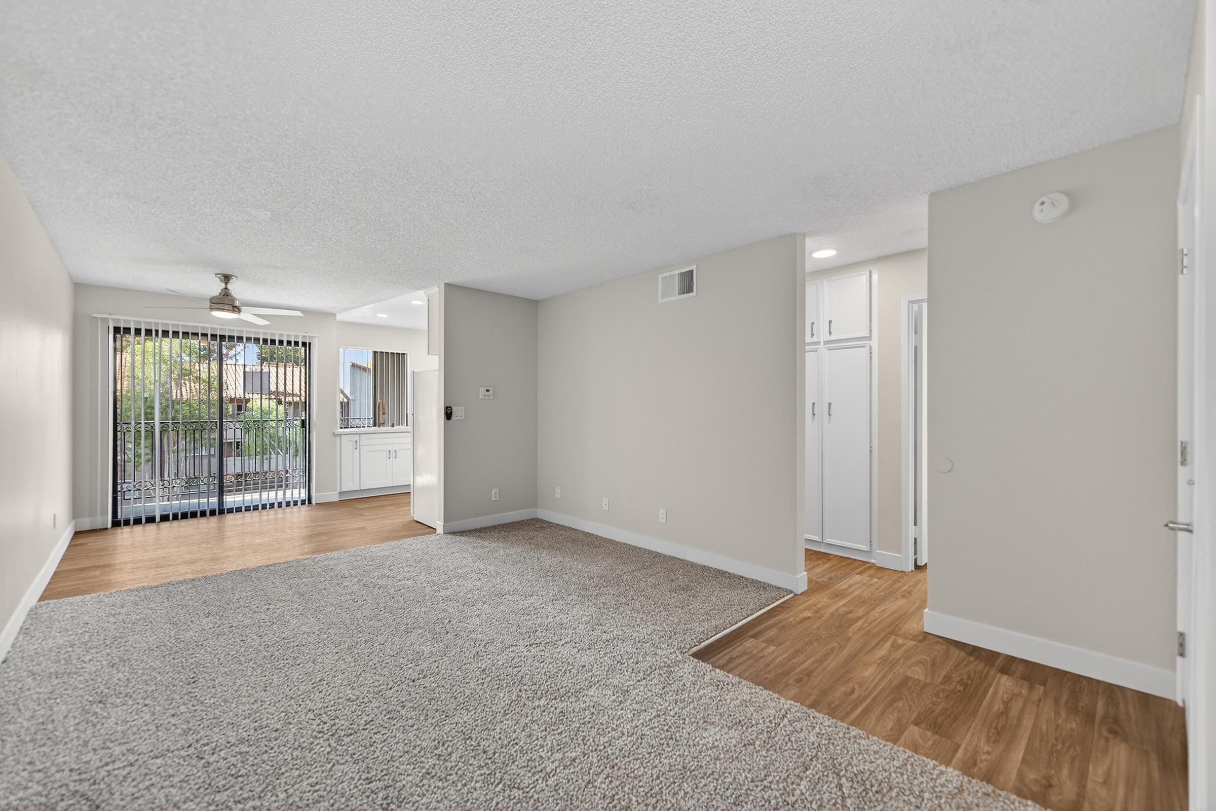 A spacious, well-lit living area featuring neutral-colored walls and carpet. On the left, a sliding glass door leads to an outdoor patio, with a kitchen visible in the background. A hallway on the right leads to additional rooms, complemented by modern lighting fixtures.