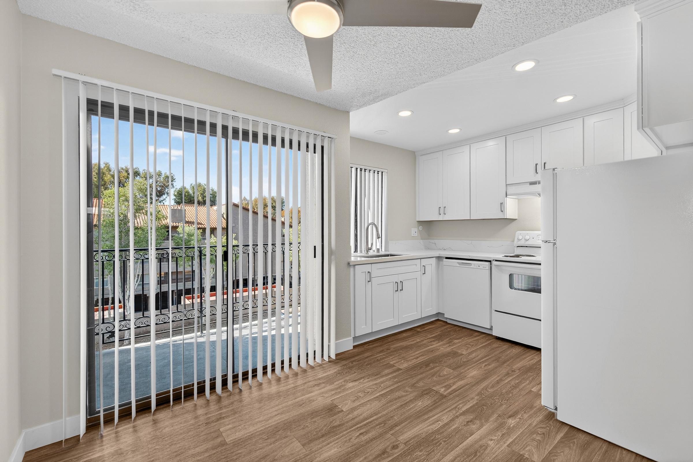 Bright kitchen with white cabinetry and appliances, featuring a ceiling fan and wood-like flooring. Large sliding glass doors lead to a balcony with views of trees and adjacent buildings. Natural light floods the space, creating a welcoming atmosphere.