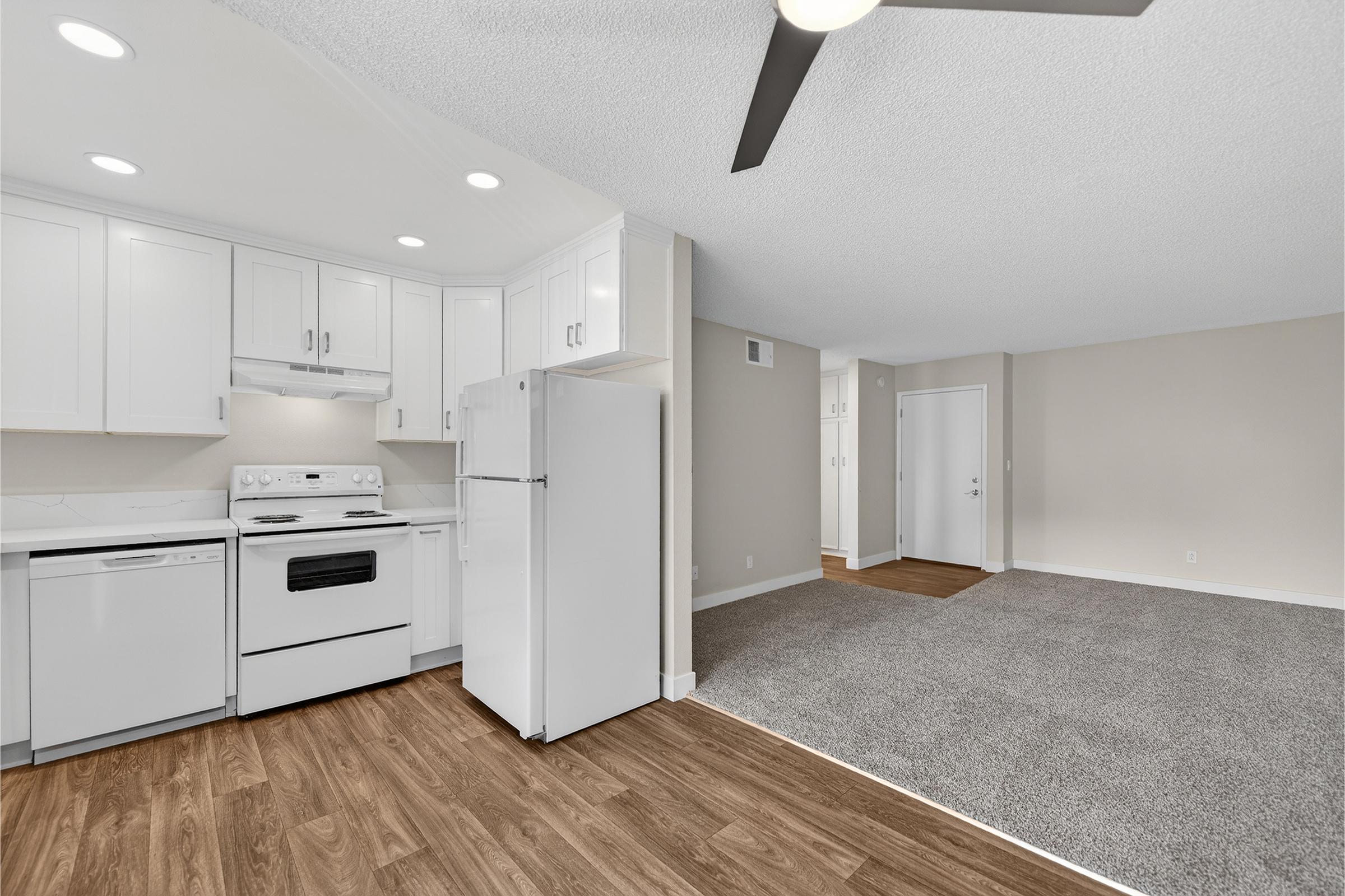Open-concept living space featuring a modern kitchen with white cabinets, a refrigerator, and a stove. The flooring is a mix of laminate and carpet. Bright lighting and a ceiling fan enhance the airy atmosphere. A doorway leads to another room, with neutral-colored walls and ample natural light.