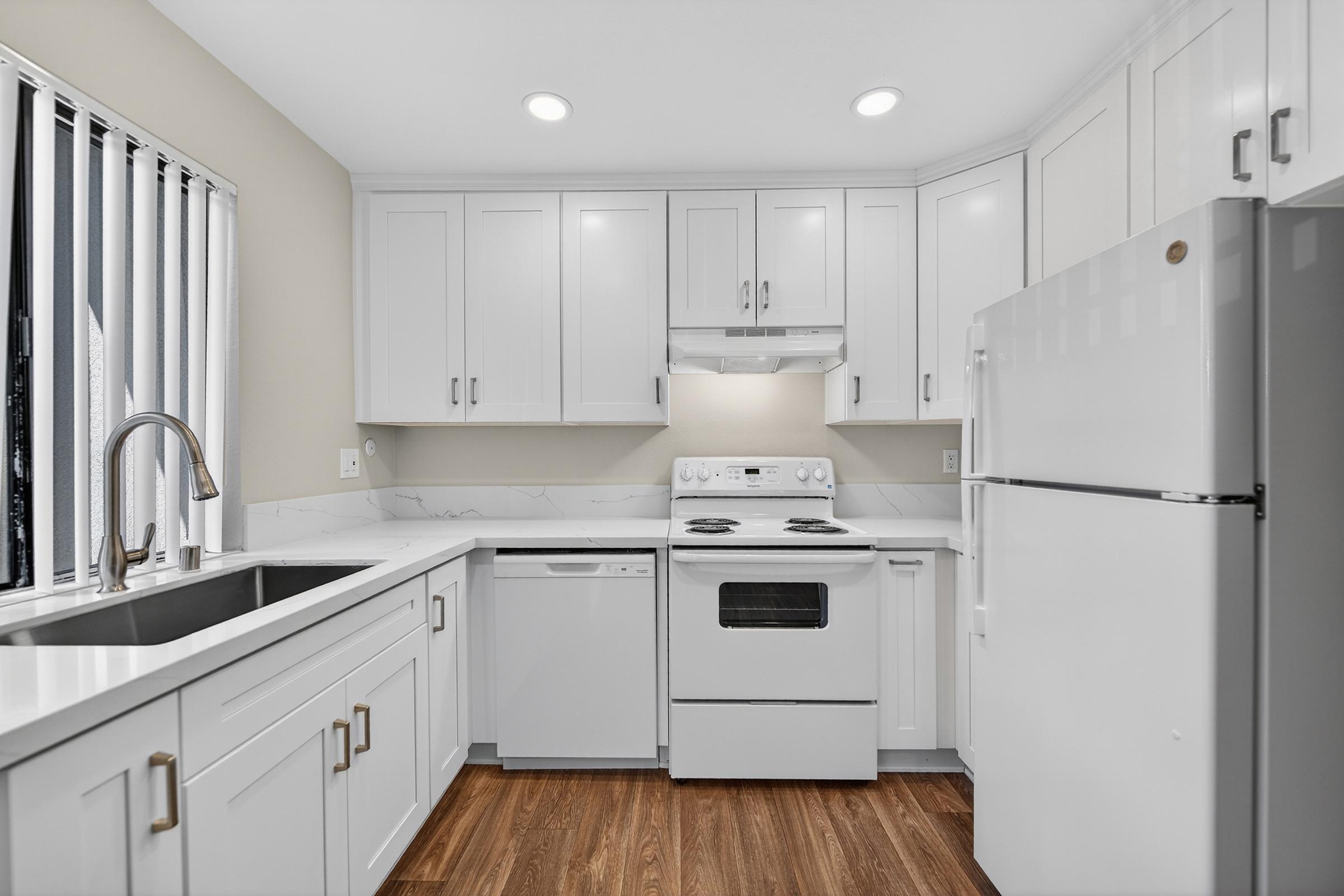 Modern kitchen featuring white cabinetry, a stainless steel sink, and a countertop with a white marble design. Appliances include a gas stove, a white refrigerator, and a dishwasher. The space is well-lit with recessed lighting and has a window with vertical blinds. The floor is made of wood.
