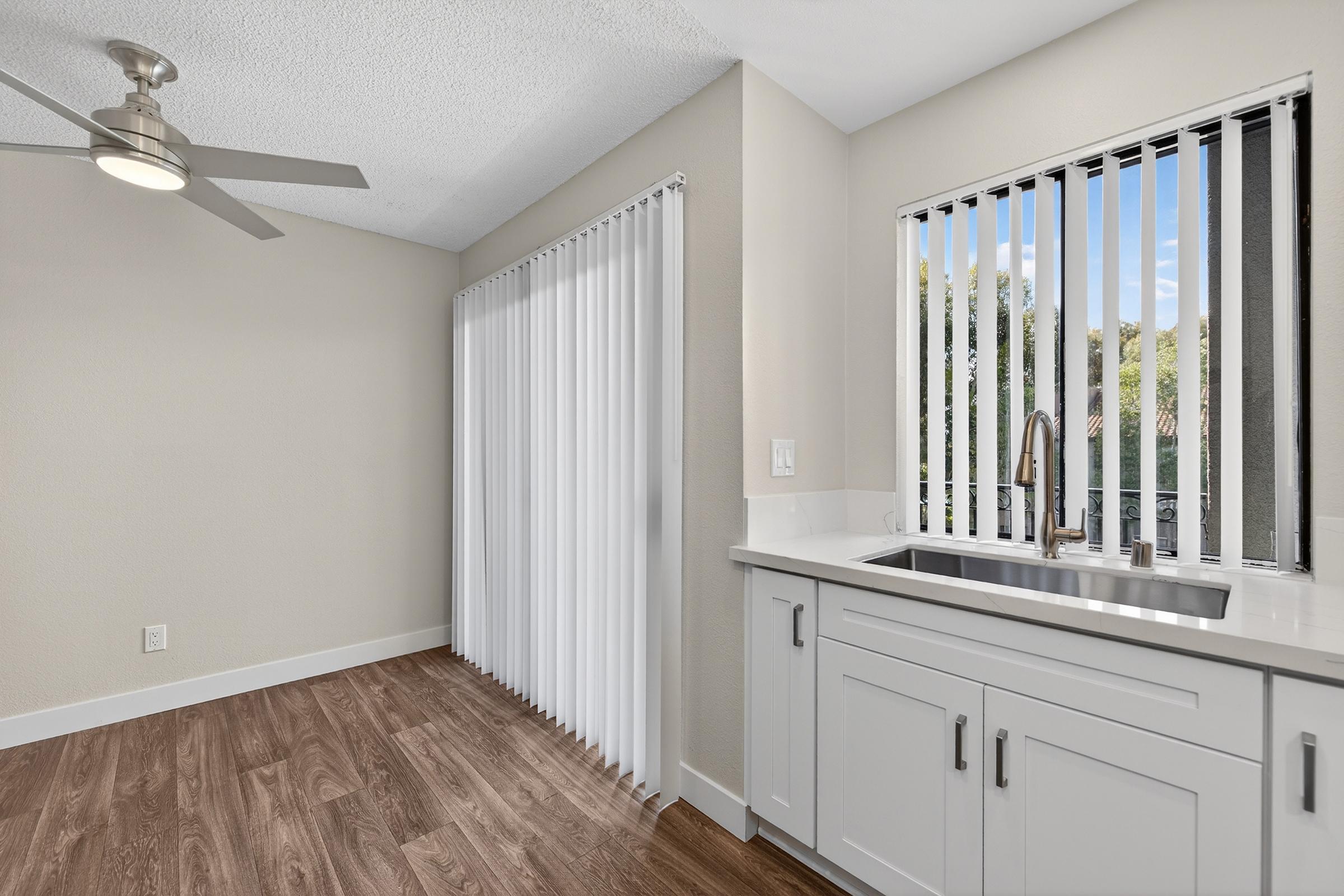 A modern kitchen with a white countertop and stainless steel sink, featuring open cabinetry. There are vertical blinds covering a window, allowing natural light to enter. The floor has wooden laminate, and a ceiling fan is visible. The walls are painted in a light, neutral color.