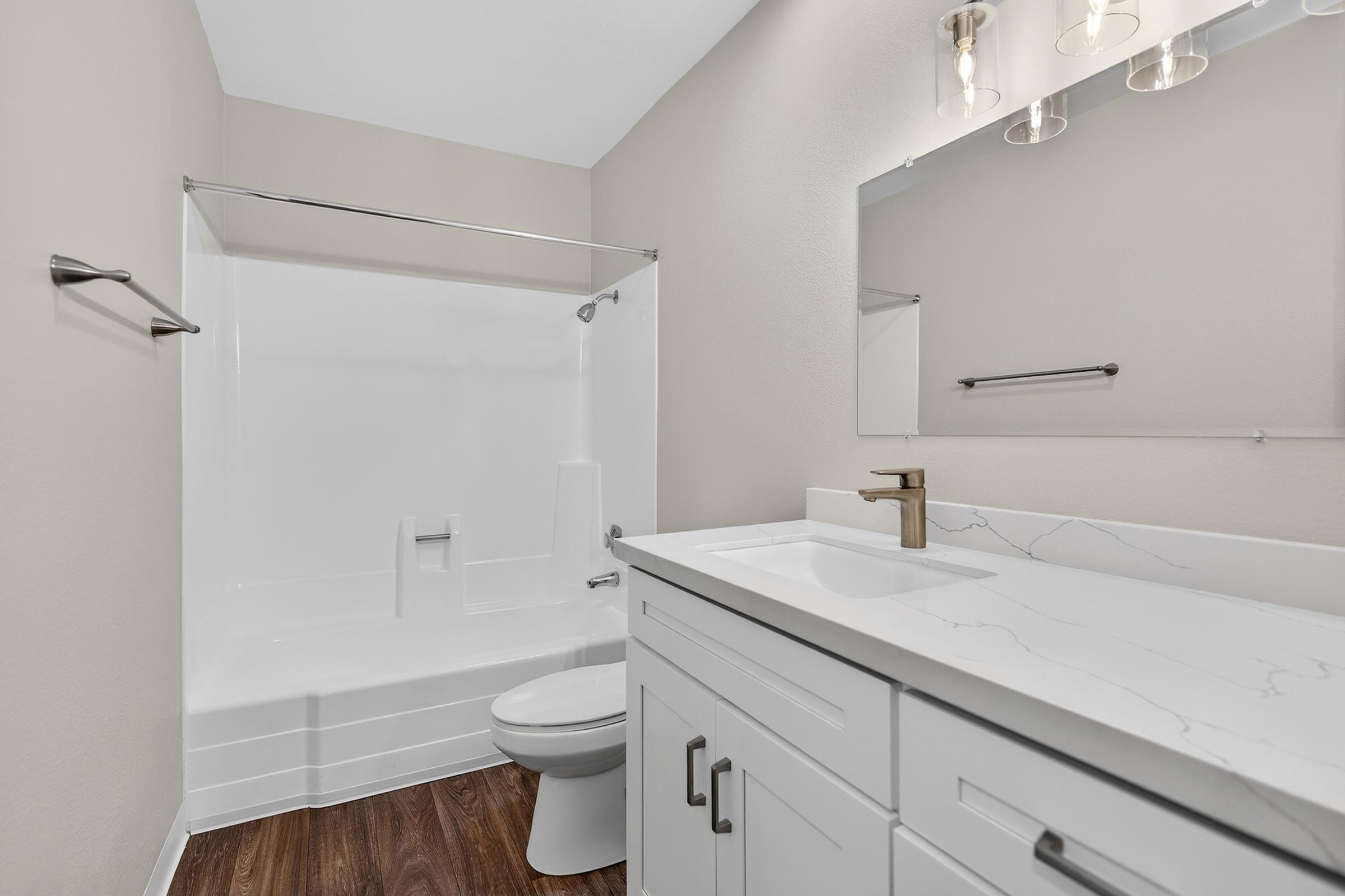 A modern bathroom featuring a white tub and shower combo, a sleek white vanity with a marble-like countertop, a gold faucet, and a large mirror. The walls are painted in a neutral tone, and the flooring is dark wood. Bright lighting fixtures illuminate the space.
