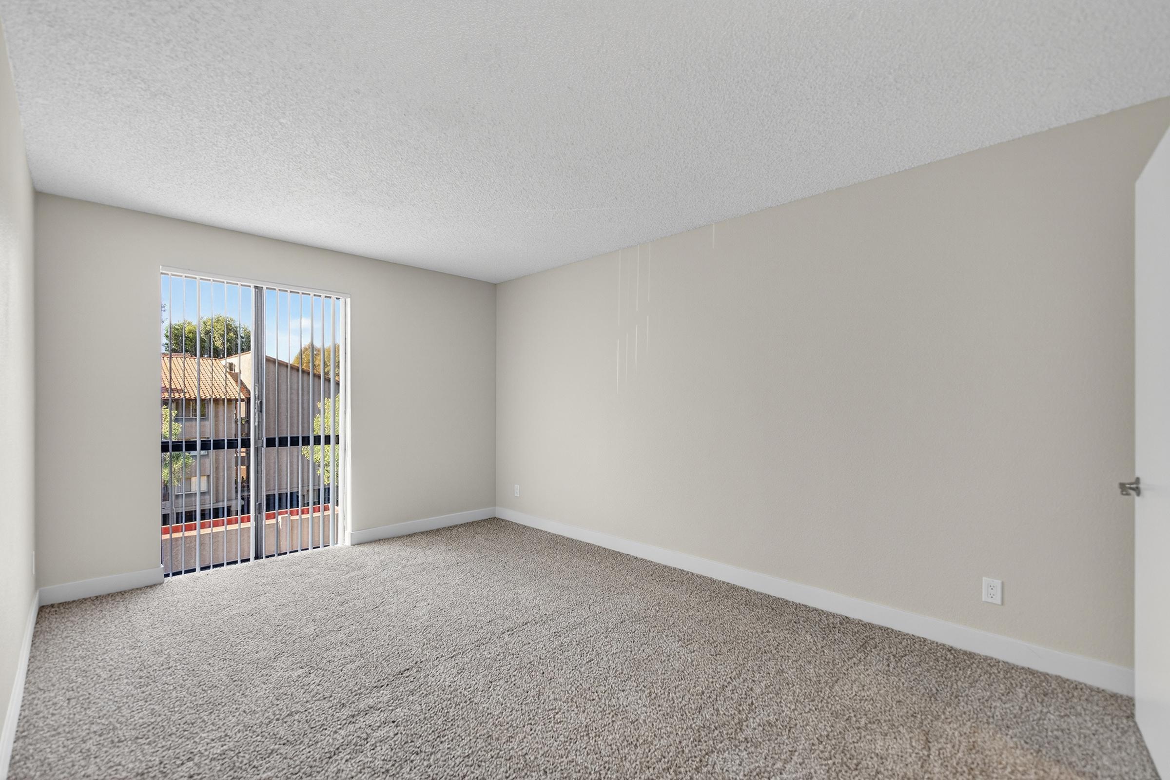 A vacant room with light beige walls and a textured ceiling. The floor is covered with plush, light gray carpet. There is a sliding glass door with vertical blinds leading to a balcony outside, allowing natural light to enter. The overall space appears clean and ready for furnishing.