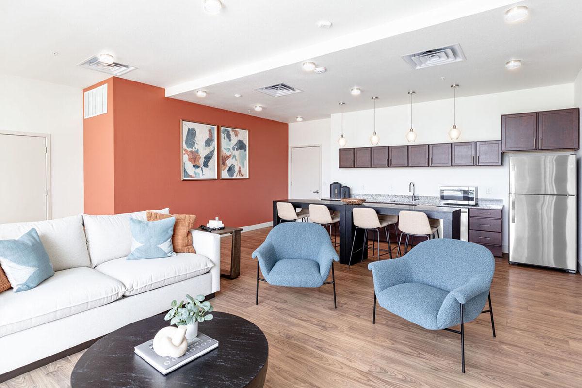 Modern lounge room featuring a light-colored sofa, two blue chairs, and a round dark coffee table. The walls are painted an accent orange, with abstract art pieces on display.