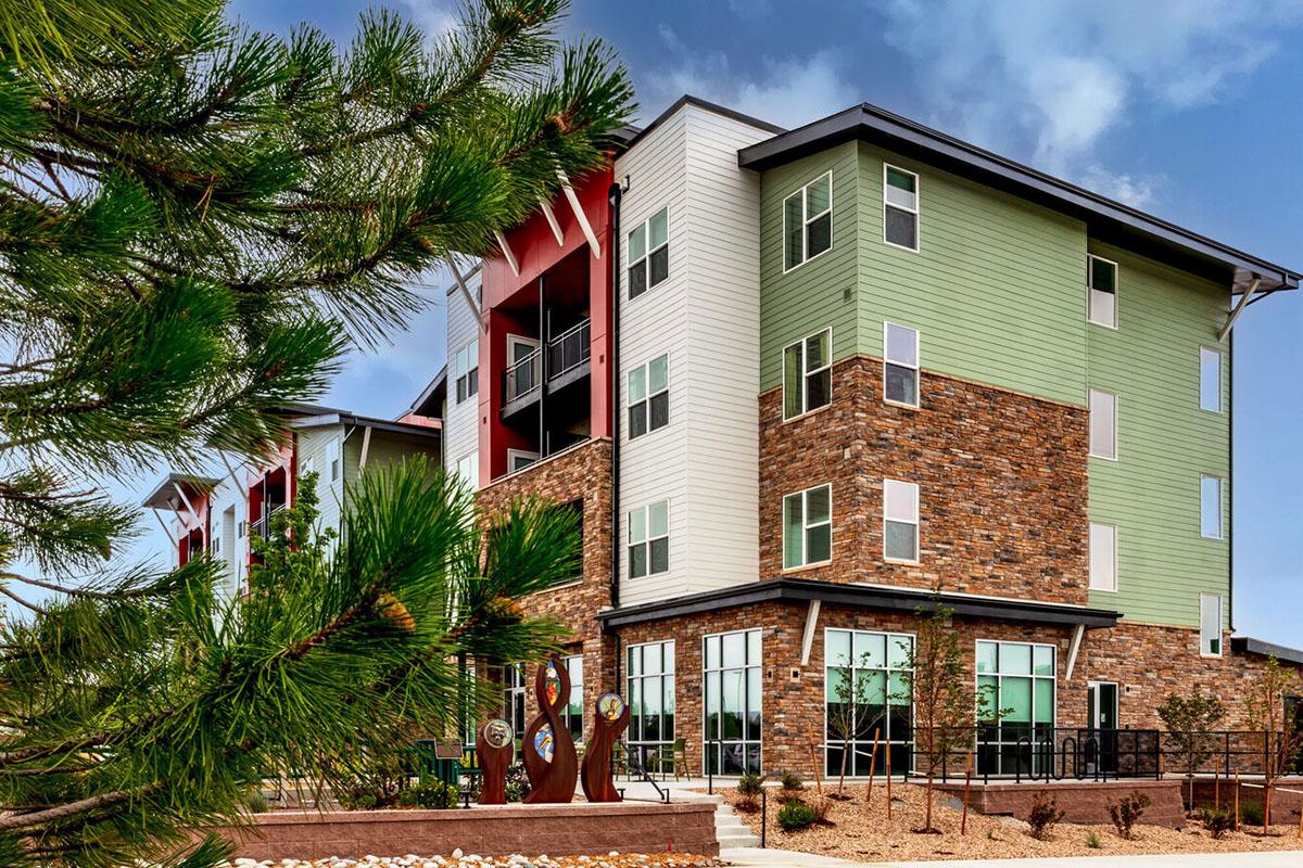 Modern apartment building featuring a mix of materials, including stone and wood. The structure has multiple stories with large windows, surrounded by landscaped greenery.