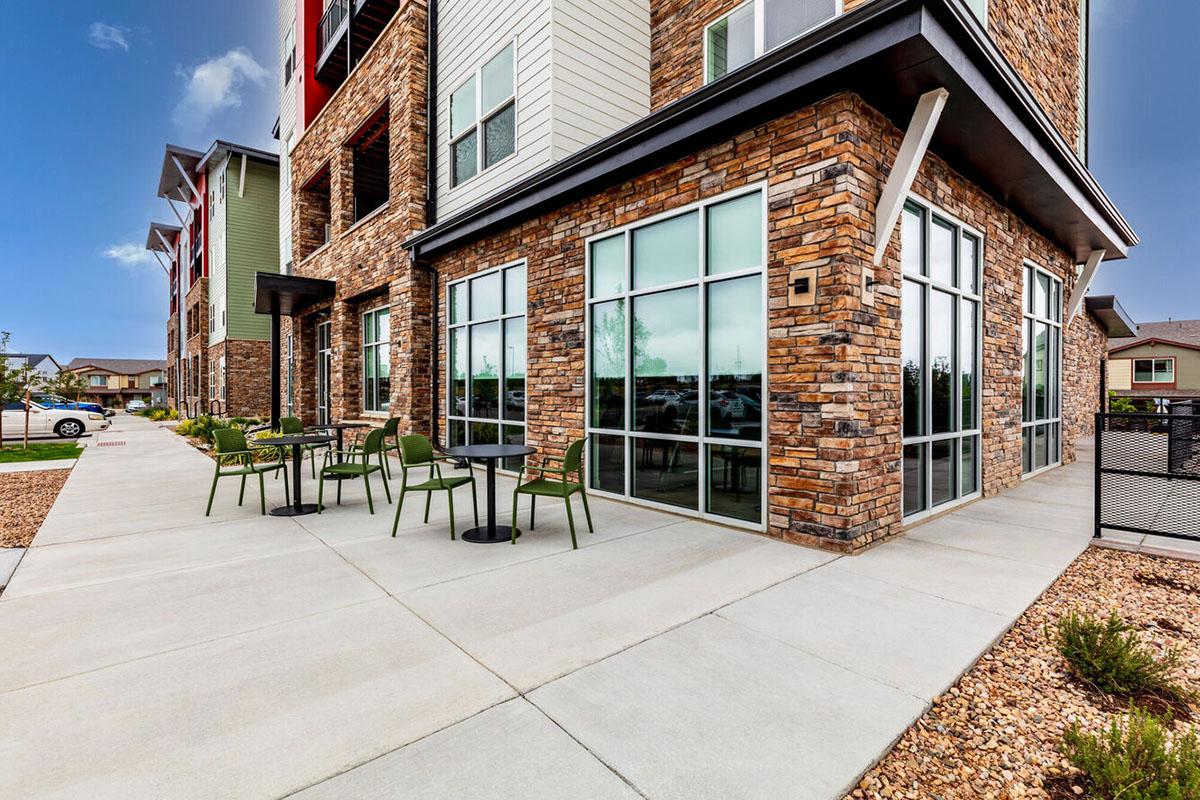 Exterior view of a modern building featuring a stone facade. In front, there are two small tables with green chairs arranged on a concrete patio.