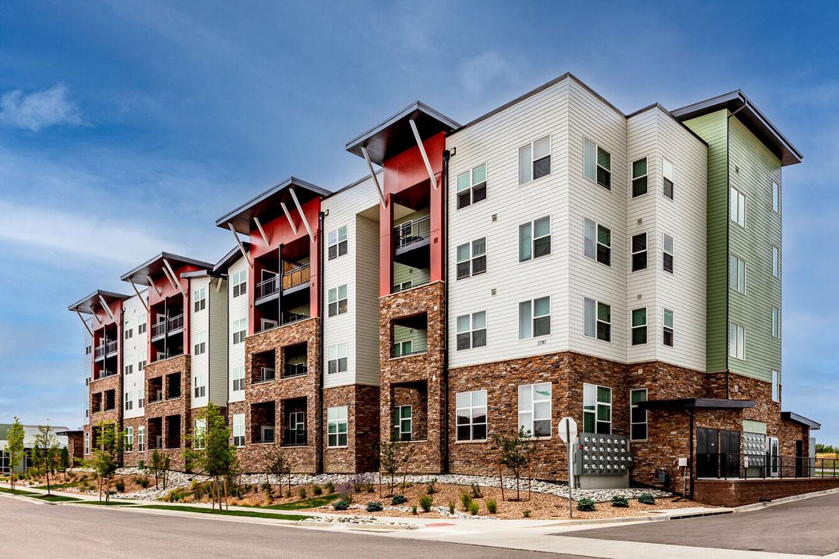 Modern apartment building with four stories featuring a mix of brick and siding exteriors. The structure has expansive windows, balconies, and a contemporary architectural design, surrounded by landscaped greenery and a well-maintained walkway.