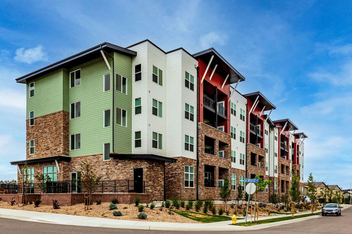 Modern multi-story apartment building with a mix of green, white, and red siding, featuring stone accents. The structure has large windows and balconies.