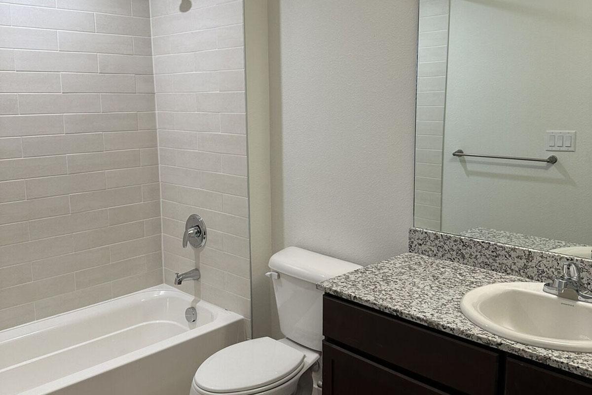A modern bathroom featuring a white bathtub and shower combination, a toilet, and a dark wood vanity with a granite countertop.