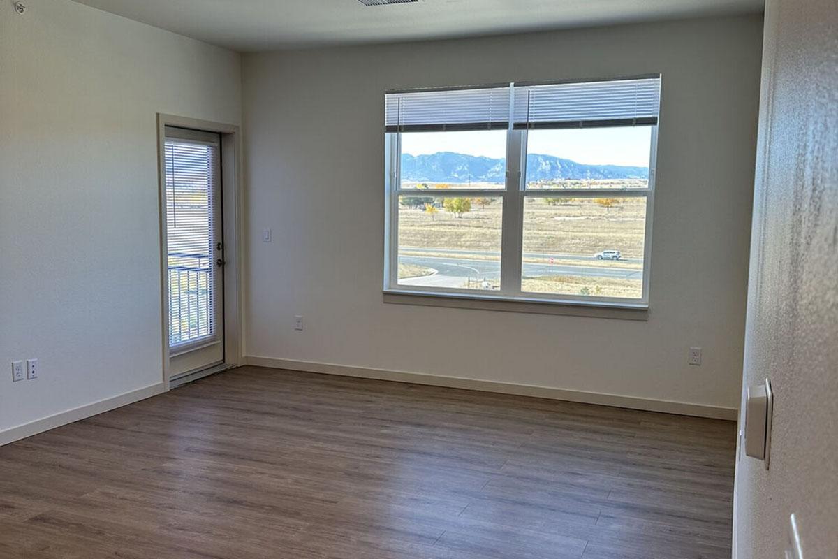 An empty room with light-colored walls and hardwood flooring. A large window lets in natural light and offers a view of mountains in the distance.
