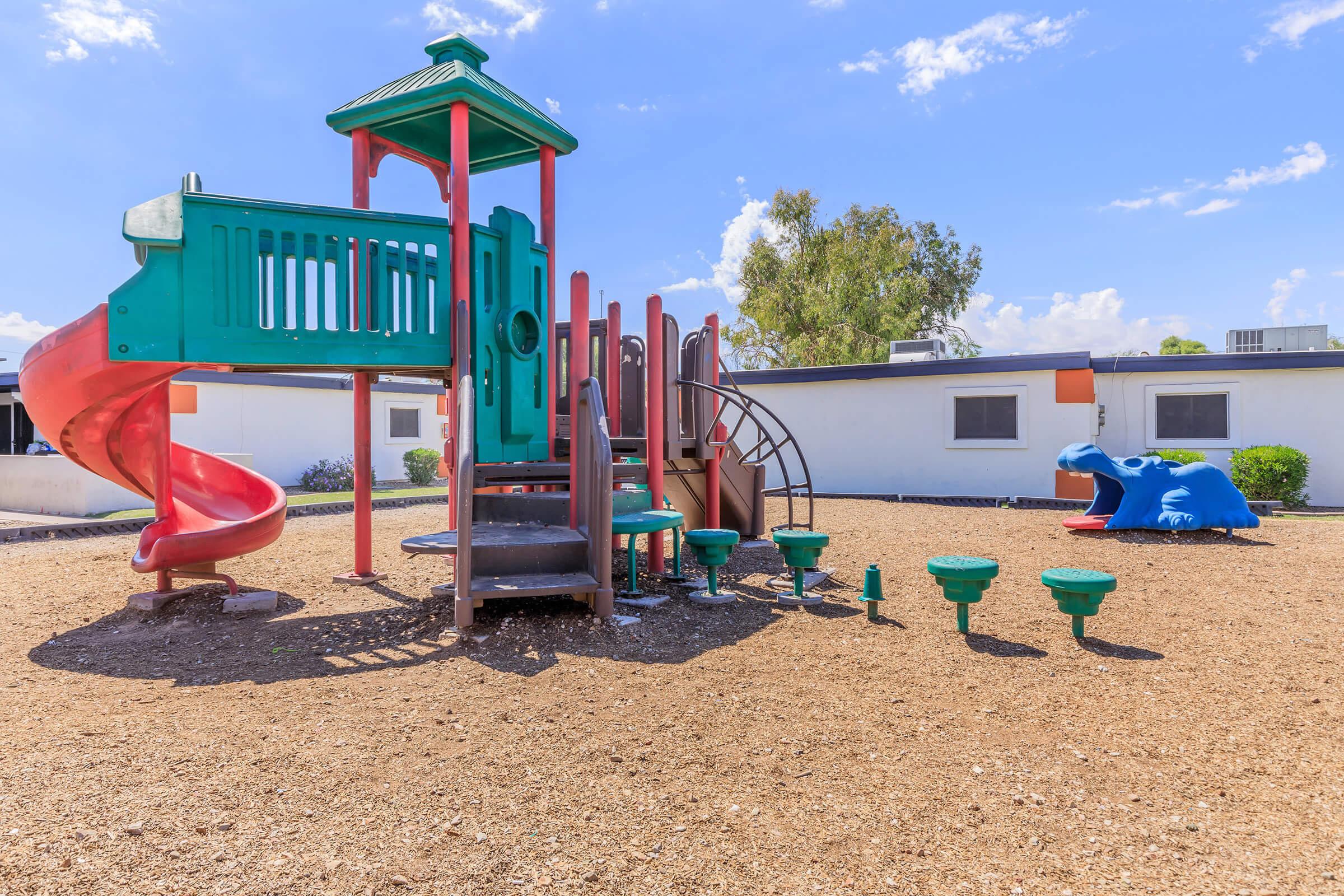 A colorful playground featuring a green and red climbing structure with a slide, situated on a sandy surface. In the background, a blue dinosaur-shaped play structure is visible. The scene is bright and sunny, with a few trees and a building in the background.