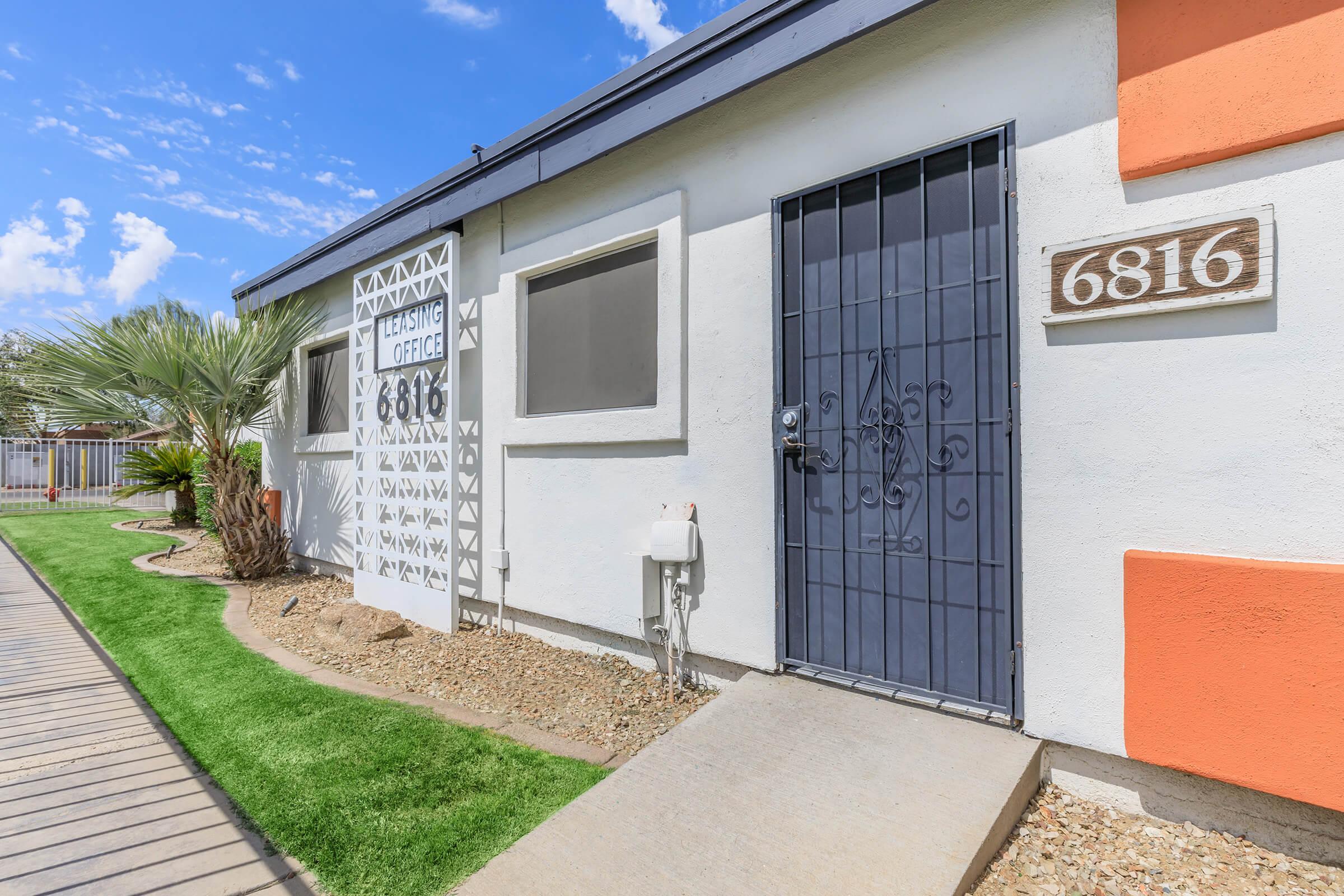 A leasing office entrance with a decorative metal gate, featuring the address "6816" on the wall. The building has a white and orange exterior, surrounded by landscaping that includes a small path, grass, and palm trees. The sky is clear with some clouds.