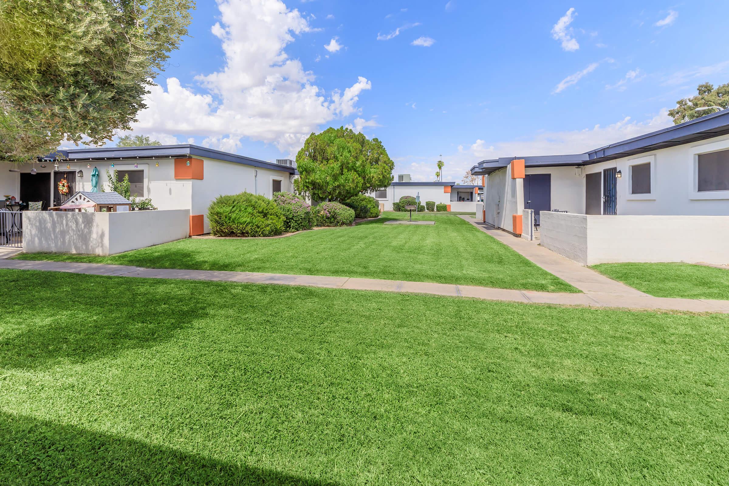 A well-maintained courtyard featuring lush green grass, bordered by modest residential buildings. The sky is bright with scattered clouds, and the pathway runs through the center, connecting the buildings on either side, creating a serene and inviting outdoor space.