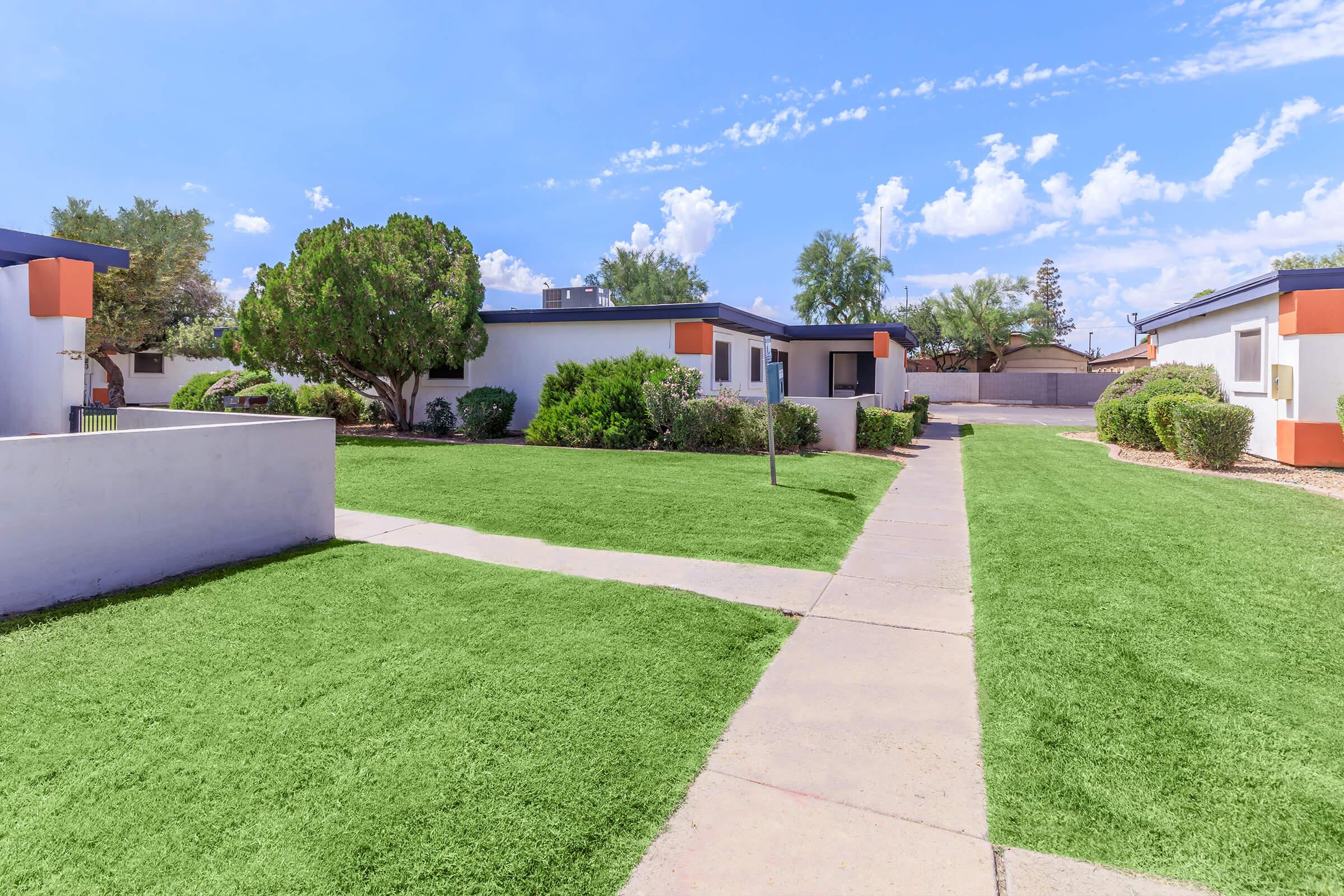 A sunny outdoor area featuring a well-maintained pathway lined with green grass, leading to a series of modern, single-story buildings. The architecture includes vibrant orange accents, surrounded by neatly trimmed shrubs and trees against a clear blue sky with a few fluffy clouds.