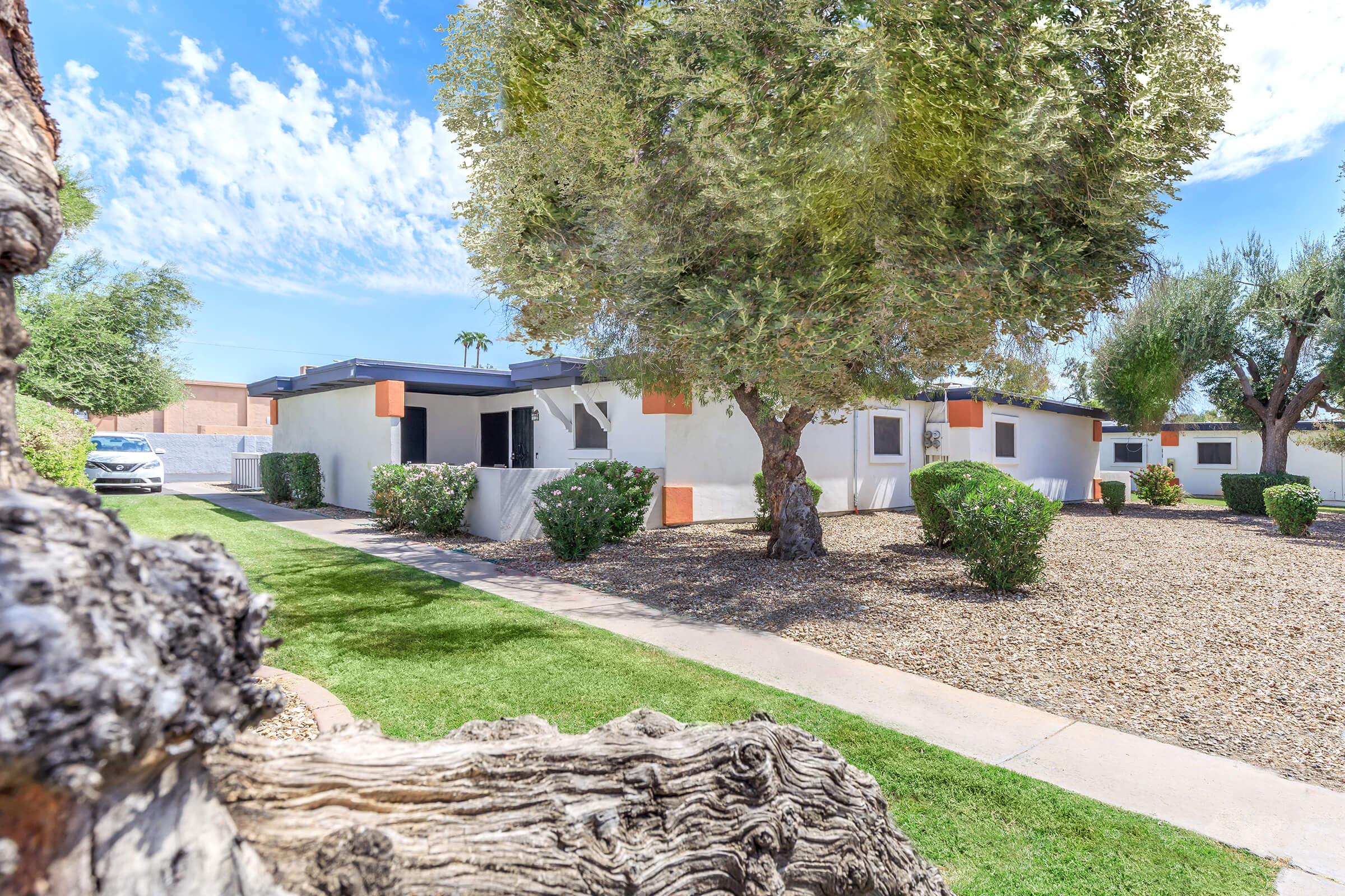 A modern single-story home surrounded by well-maintained landscaping, featuring lush green grass and mature trees. The exterior is painted in white with orange accents. A gravel area and a pathway lead to the entrance, while a car is visible in the background under a bright blue sky with scattered clouds.