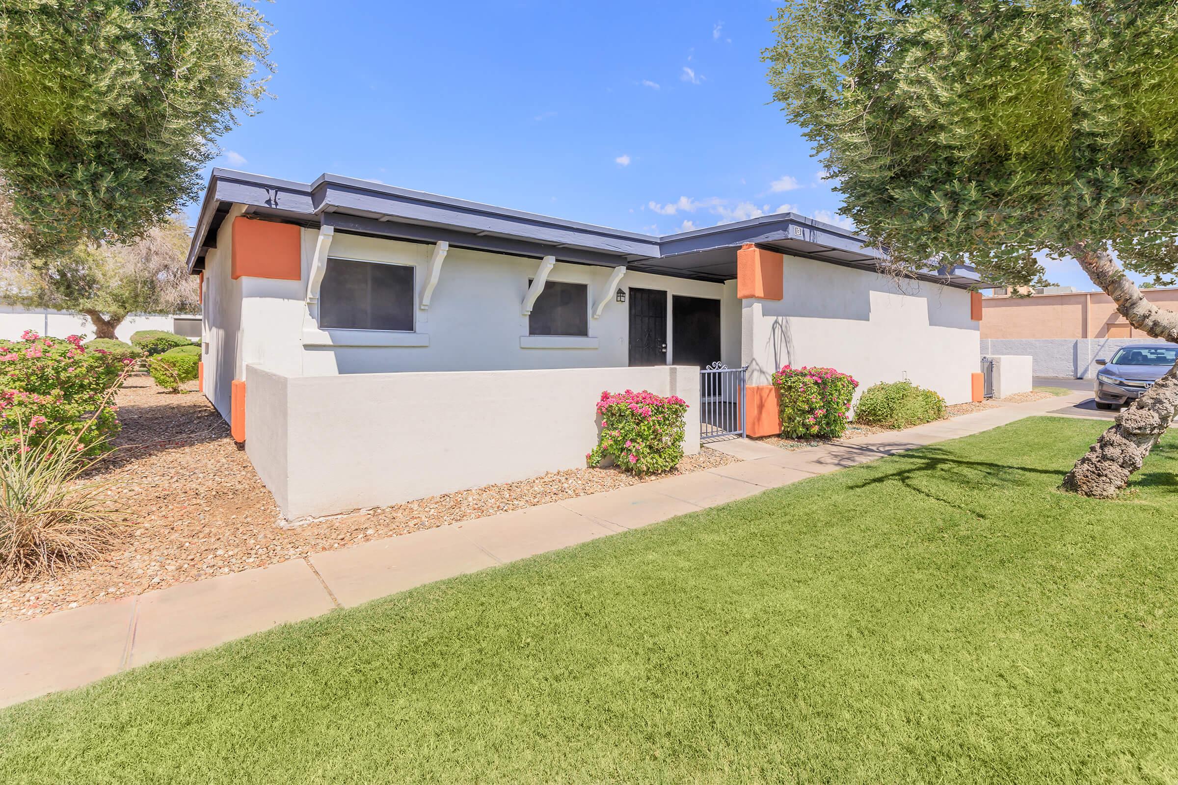 A single-story residential building with a light-colored exterior and orange accents. The property features a well-manicured lawn, shrubs, and small trees. A pathway leads to the entrance, and there are flower beds on either side, creating an inviting outdoor space. Clear blue sky in the background.