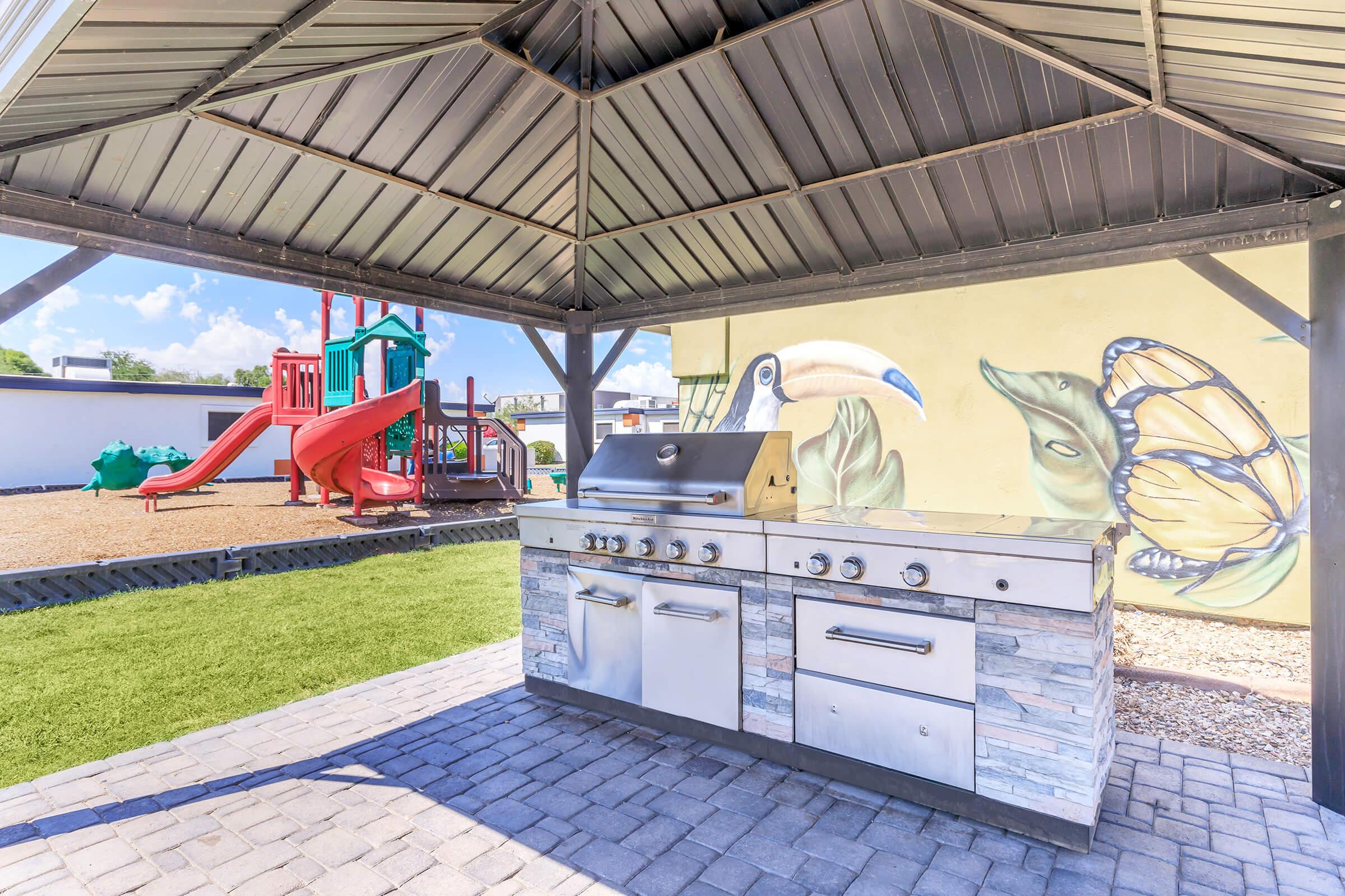 Outdoor barbecue grill under a shelter with a metal roof. The grill features various cooking stations. In the background, there is a colorful playground with slides and climbing structures, adjacent to a wall featuring a mural of a bird and butterfly. The area has plush green grass and a stone-paved surface.