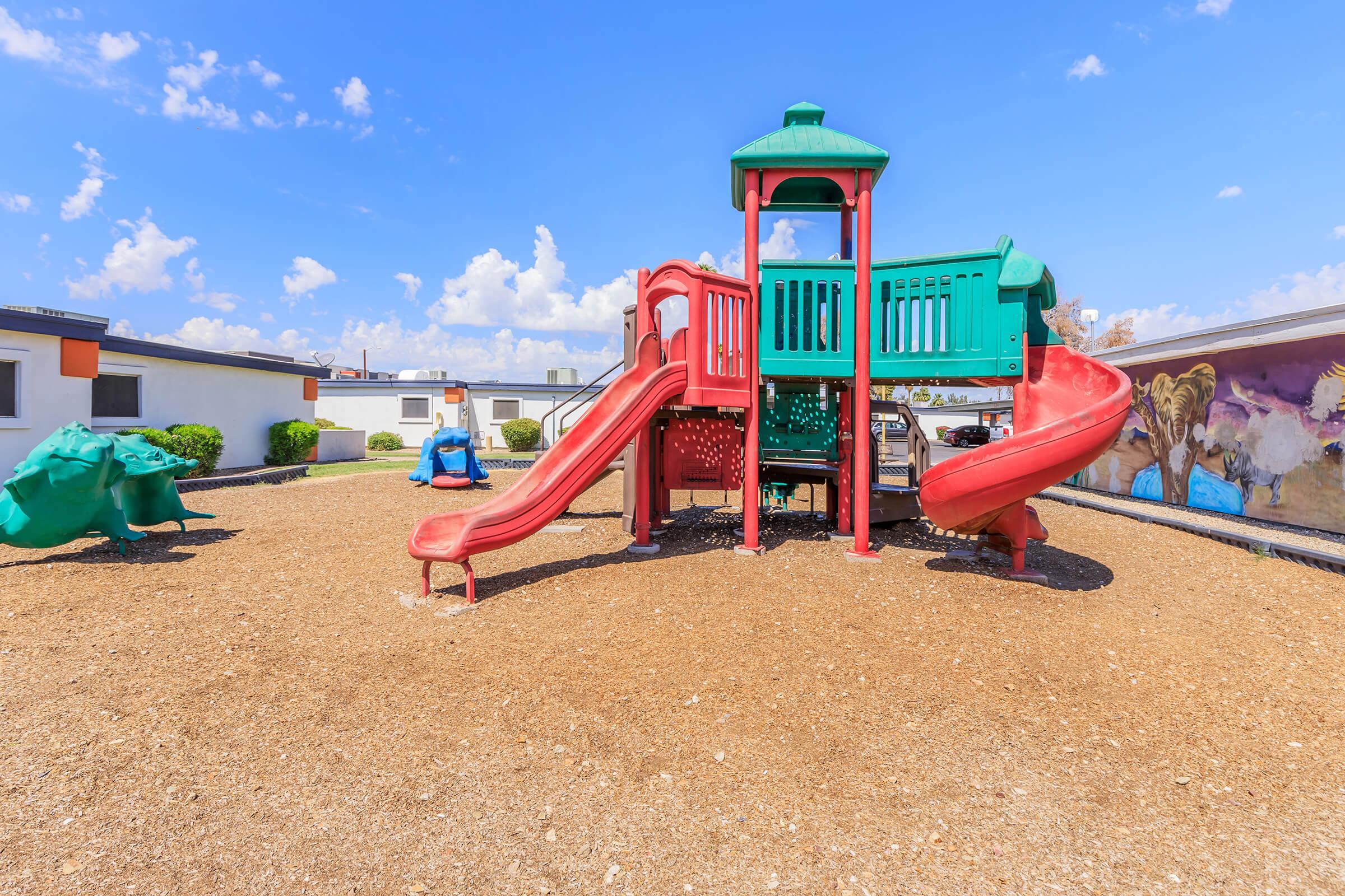 A colorful playground with a red and green play structure featuring slides and climbing areas, set on a sandy surface. In the background, there are buildings and a mural, with a clear blue sky dotted with clouds. Empty swings are visible, indicating a playful atmosphere.