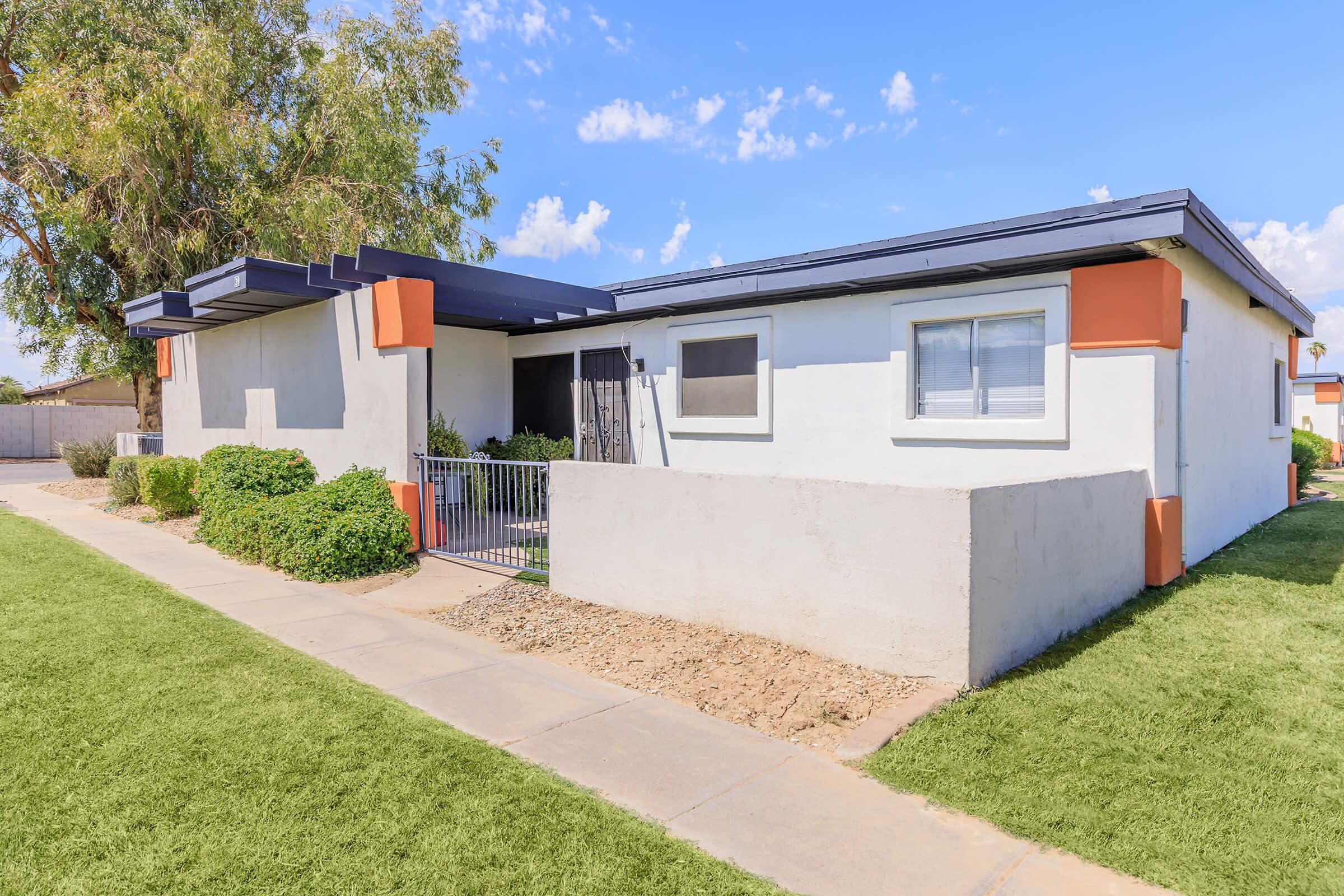 Modern single-story house with a light-colored exterior and orange accents, surrounded by neatly trimmed grass and shrubs. A concrete pathway leads to the entrance, which features a gate. Clear blue sky and a few clouds are visible in the background.