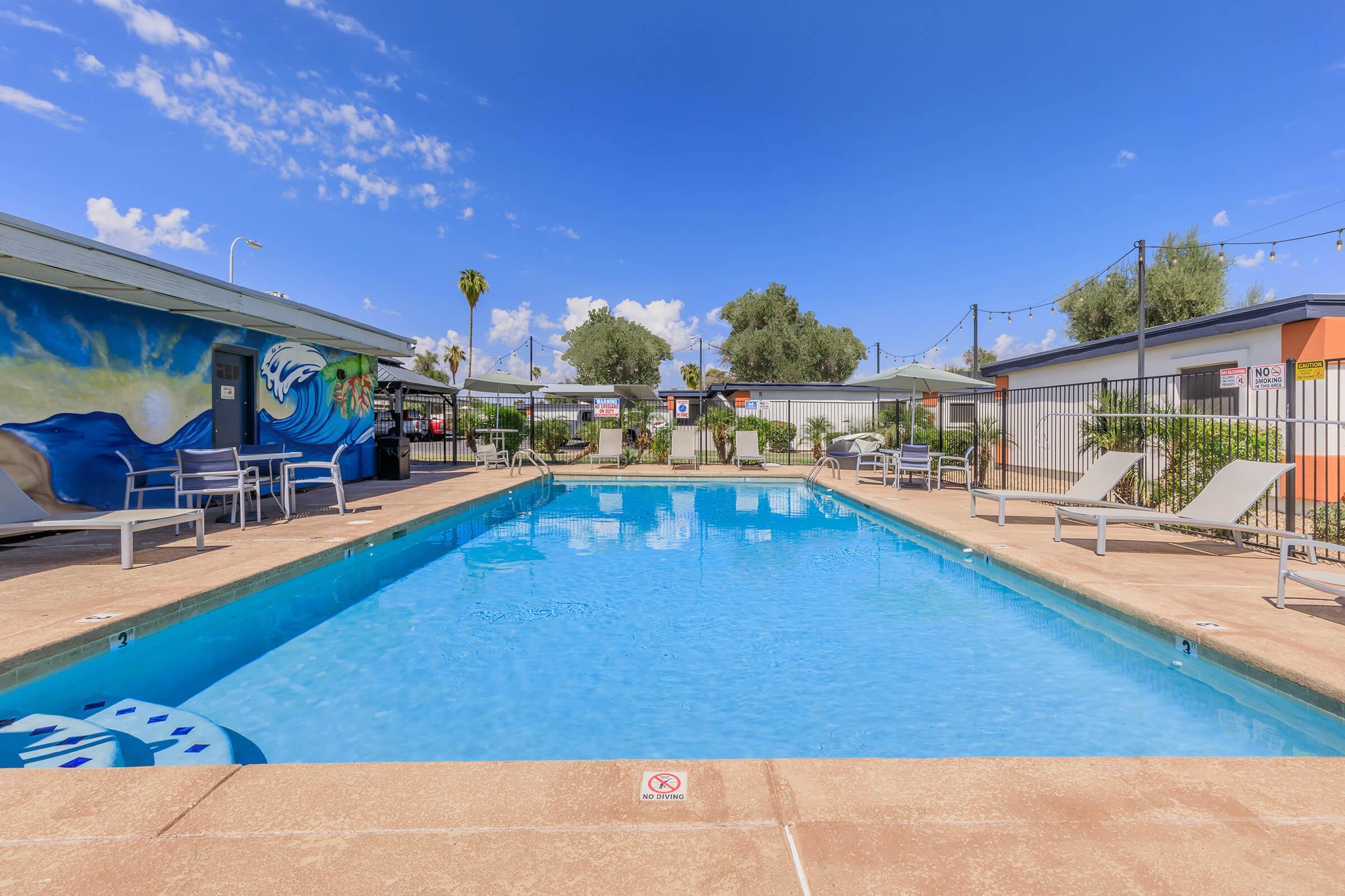 A clear blue swimming pool surrounded by lounge chairs and a vibrant mural on the wall. The sky is bright with fluffy clouds, and palm trees are visible in the background. There are signs around the pool area, adding to the welcoming atmosphere of the space.