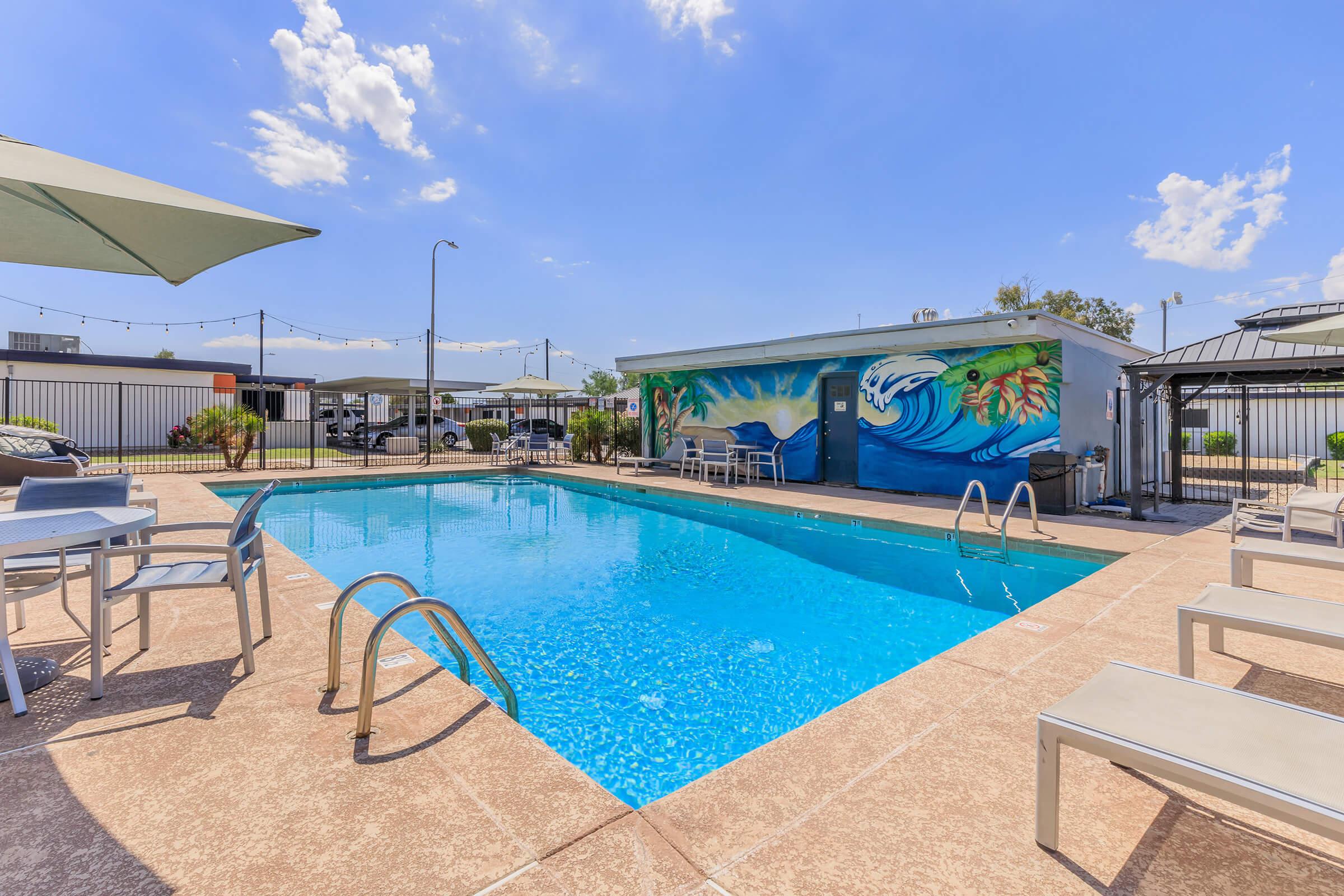 A clear blue swimming pool surrounded by lounge chairs and umbrellas. A colorful mural featuring ocean waves and sea life is on the wall near the pool. Bright blue sky with scattered clouds above, and a well-maintained outdoor area in the background.