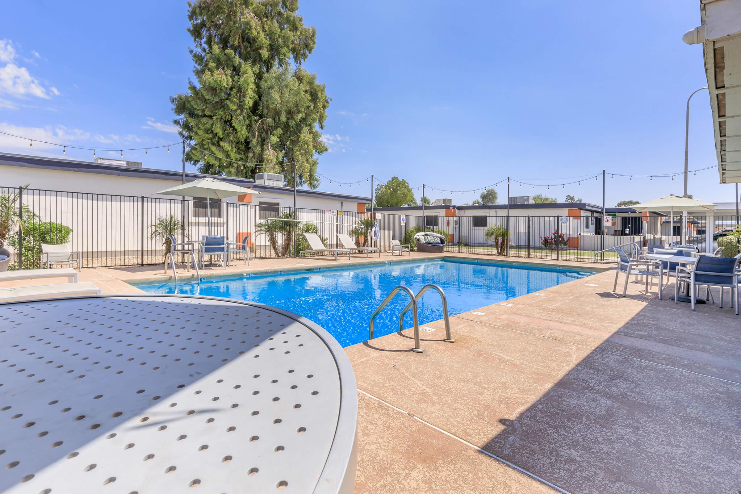 A clear blue swimming pool surrounded by lounge chairs and umbrellas. Nearby, there are potted plants and a few light fixtures strung overhead, creating a cozy atmosphere. In the background, there are several single-story buildings with a tidy layout. The sky is bright and partly cloudy.