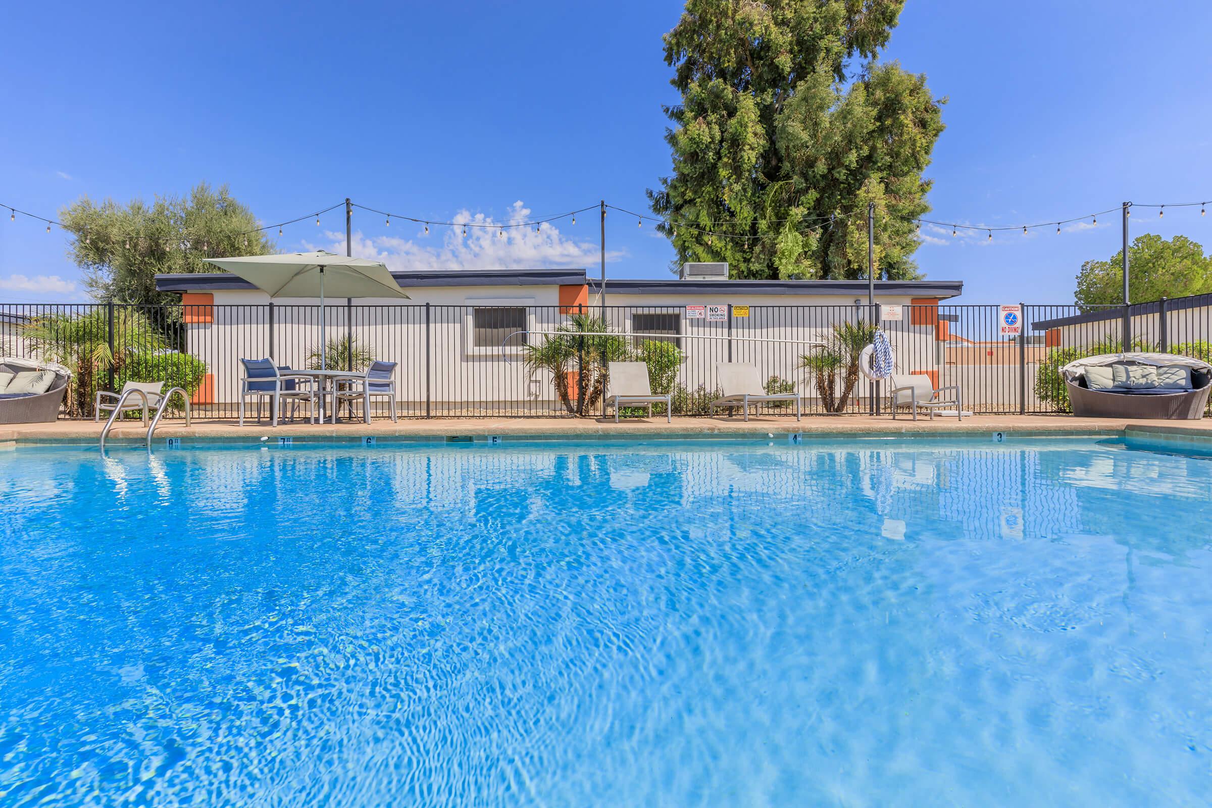 A clear blue swimming pool with inviting water, surrounded by a fence. In the background, there's a modern building with outdoor seating areas and greenery. The scene is set under a bright blue sky, adding to the serene atmosphere. String lights are visible, enhancing the ambiance for evening use.
