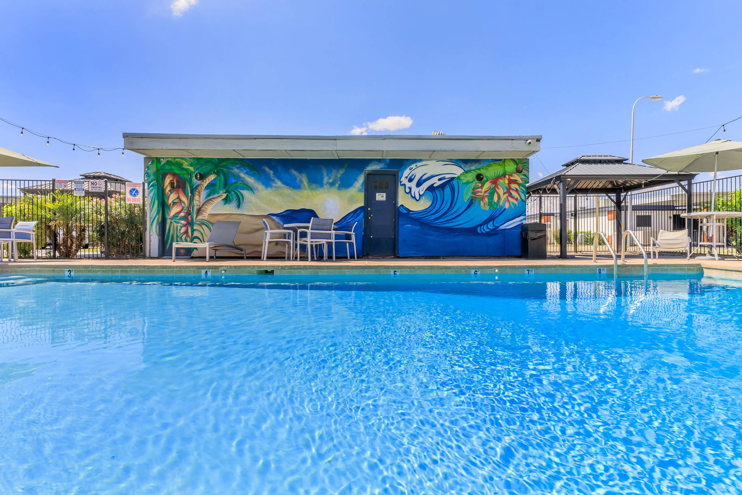 A vibrant pool area featuring a clear blue pool, surrounded by lounge chairs. In the background, there's a colorful mural depicting ocean waves and tropical plants, with a building structure and umbrellas visible under a bright sky.