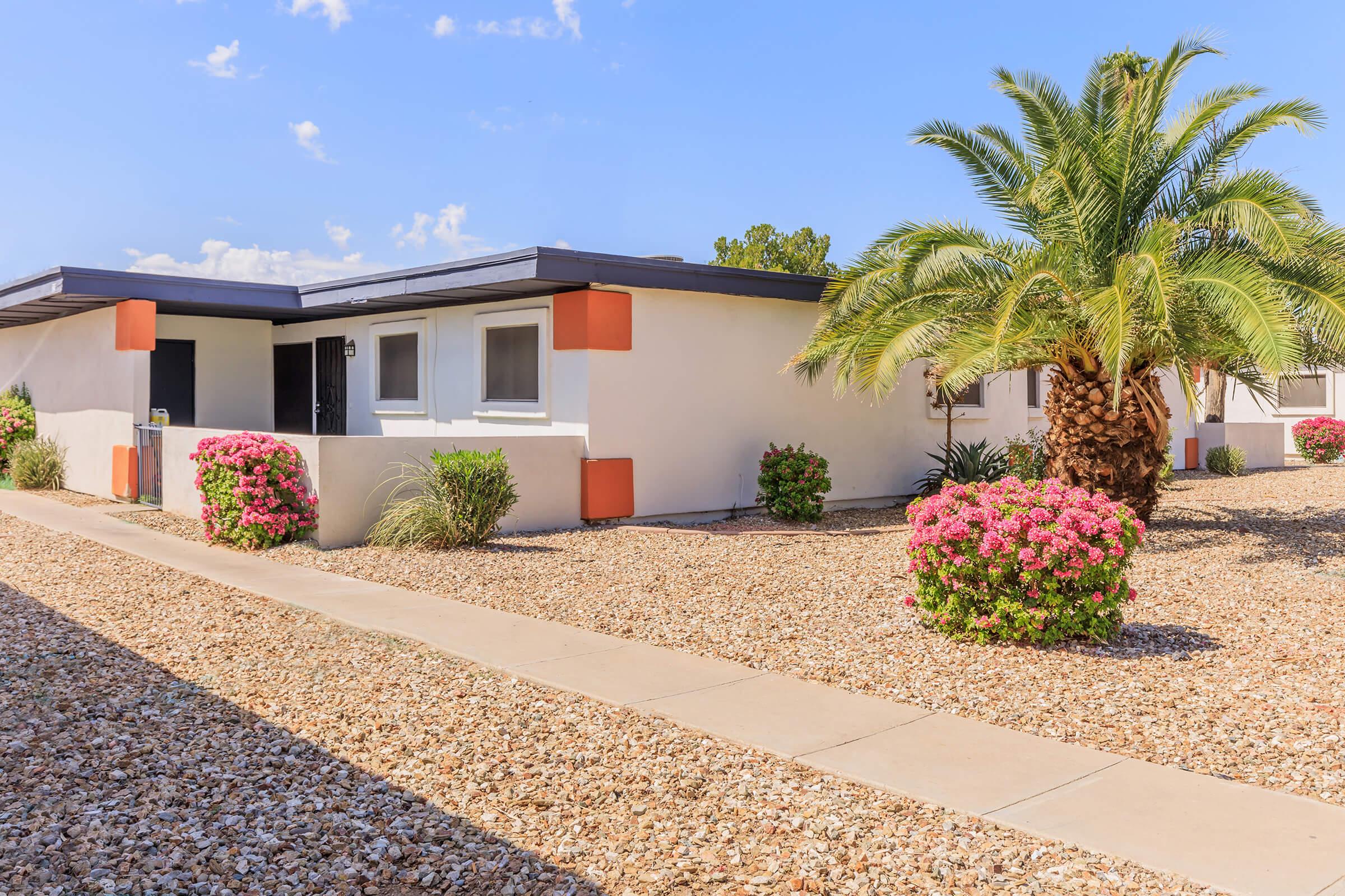 A desert-style home front with a landscaped yard featuring flowering bushes and a palm tree. The pathway leads to a single-story building with light-colored walls and orange accents. Clear blue skies create a bright, sunny atmosphere.