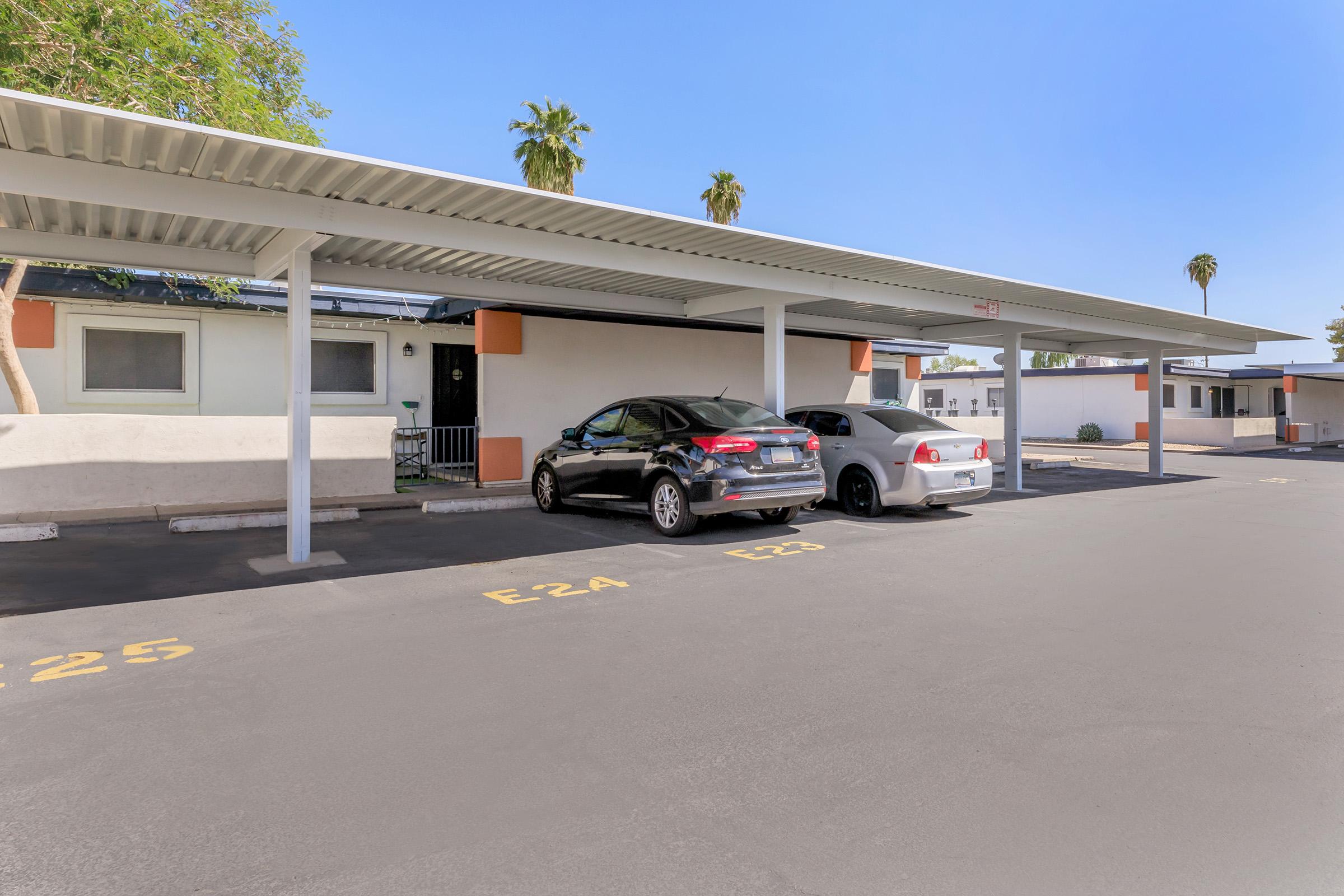 A carport area with two parked cars: a black sedan and a white sedan. The background features several palm trees and a low-rise building with a light-colored facade. The parking spaces are labeled with numbers, and there is clear blue sky above.