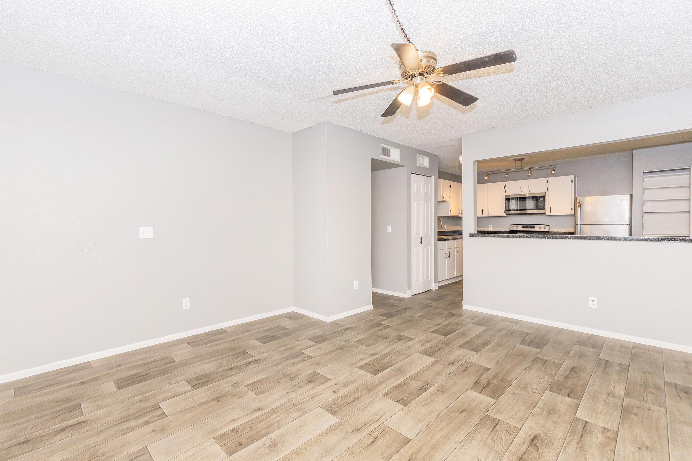 A spacious living area featuring light gray walls and a light wood-look floor. A ceiling fan hangs from the textured ceiling. In the background, a doorway leads to a kitchen with modern appliances and cabinetry. The space appears clean and inviting, ideal for comfortable living.