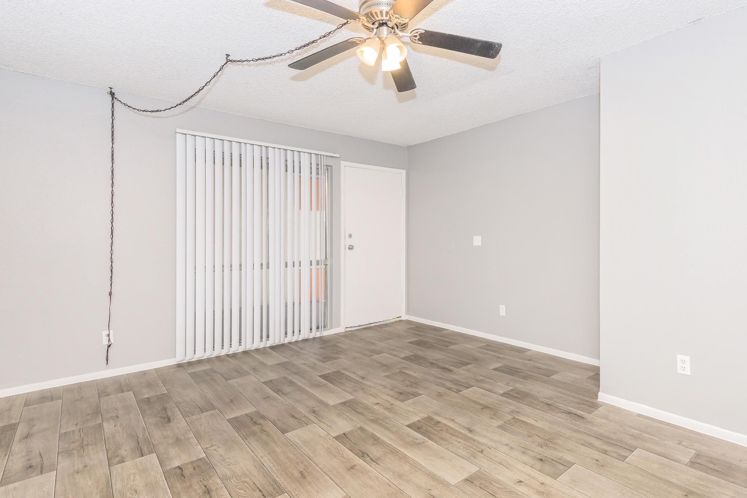A spacious room with light gray walls and wooden flooring. There is a ceiling fan with five blades and a light fixture. A set of vertical blinds covers a window, and a door is visible to the right. The overall space appears bright and uncluttered, suitable for various interior setups.